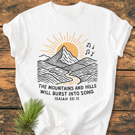 Mountains And Hills - Vintage Wash Tee features a mountain and sun graphic with musical notes and Isaiah 55:12 text, laid on wood with a pine sprig accentuating its rustic charm.