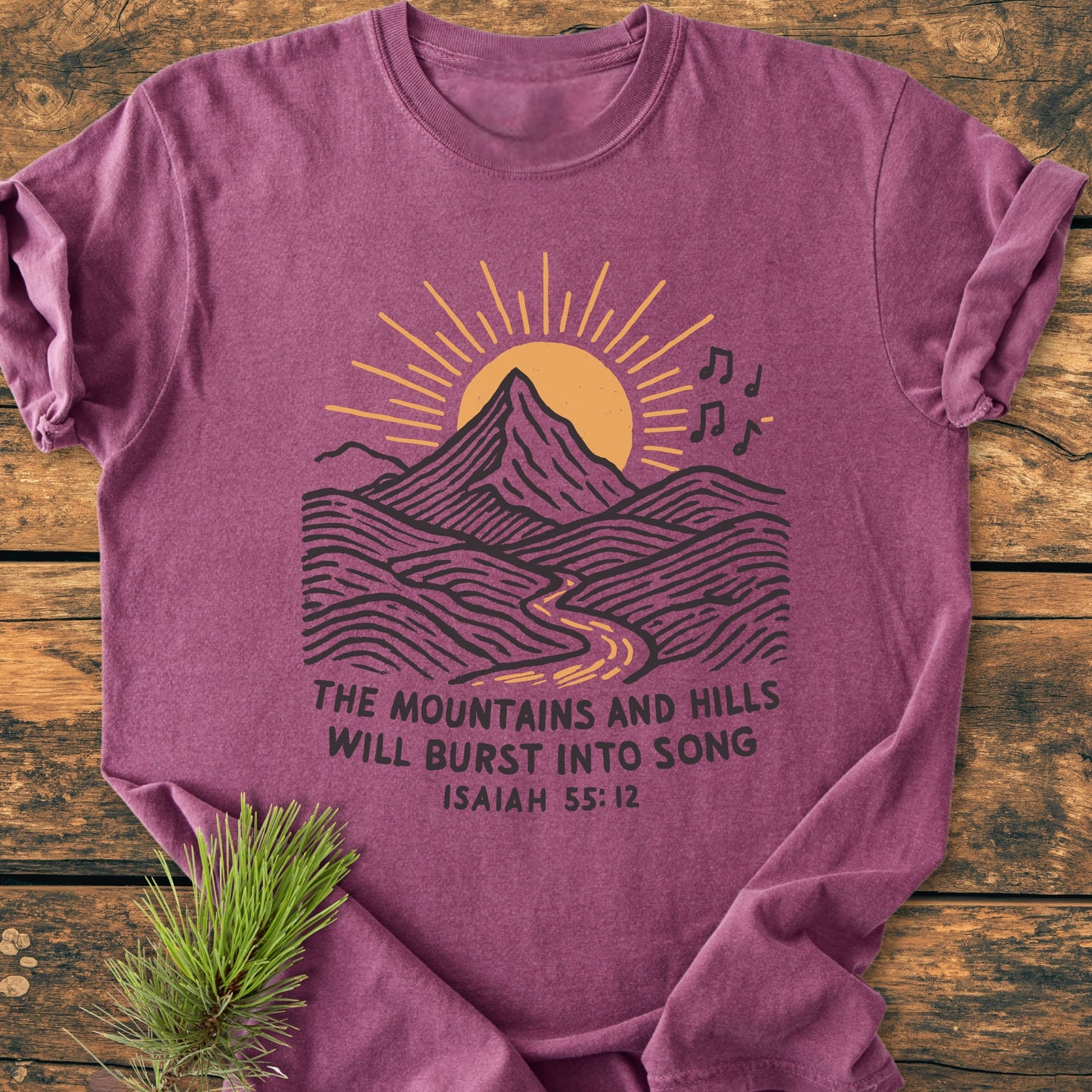Mountains And Hills - Vintage Wash Tee: Maroon T-shirt with mountain and sun graphic, Bible verse Isaiah 55:12, laid on wood, pine sprig beside. Comfortable, 100% cotton, relaxed fit, durable stitching.