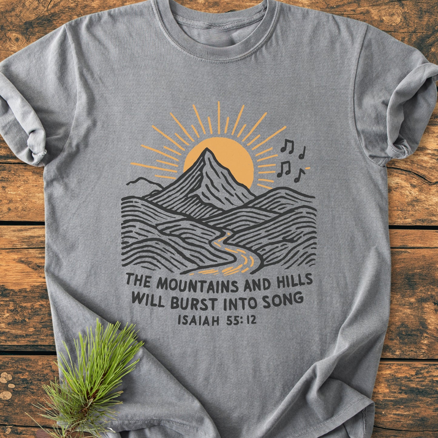 Mountains And Hills - Vintage Wash Tee: Heather-gray T-shirt with mountain landscape, golden sun, musical notes, and Bible verse. Laid on wood with pine sprig, highlighting rustic charm and comfortable fit.