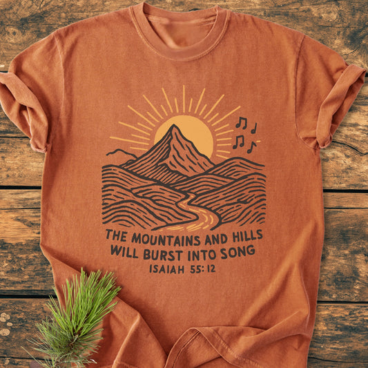 Mountains And Hills - Vintage Wash Tee features a line drawing of mountains and musical notes with Isaiah 55:12 text, laid on wood. The garment-dyed rust-orange cotton ensures comfort and durability.