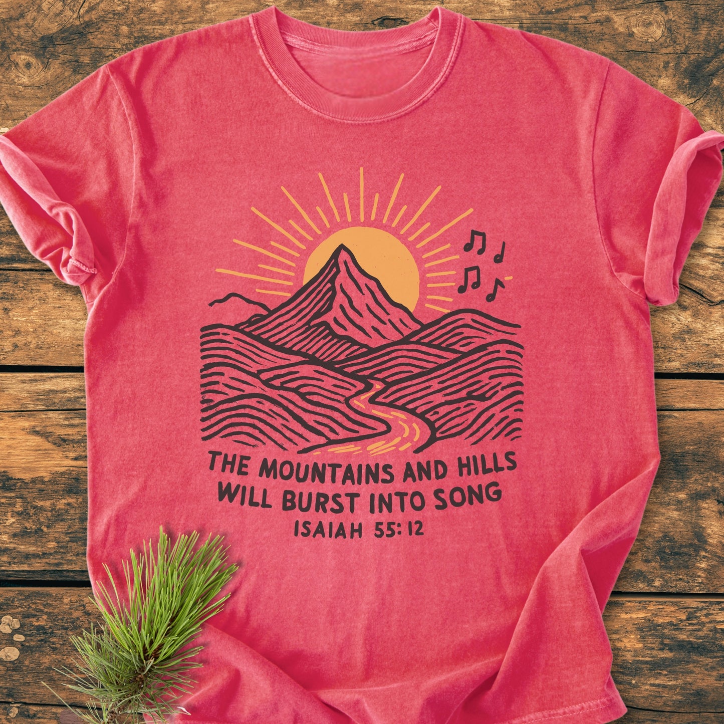 Mountains And Hills - Vintage Wash Tee featuring a stylized landscape graphic and biblical text, laid flat on a wooden surface with a pine sprig beside it, showcasing its relaxed fit and durability.