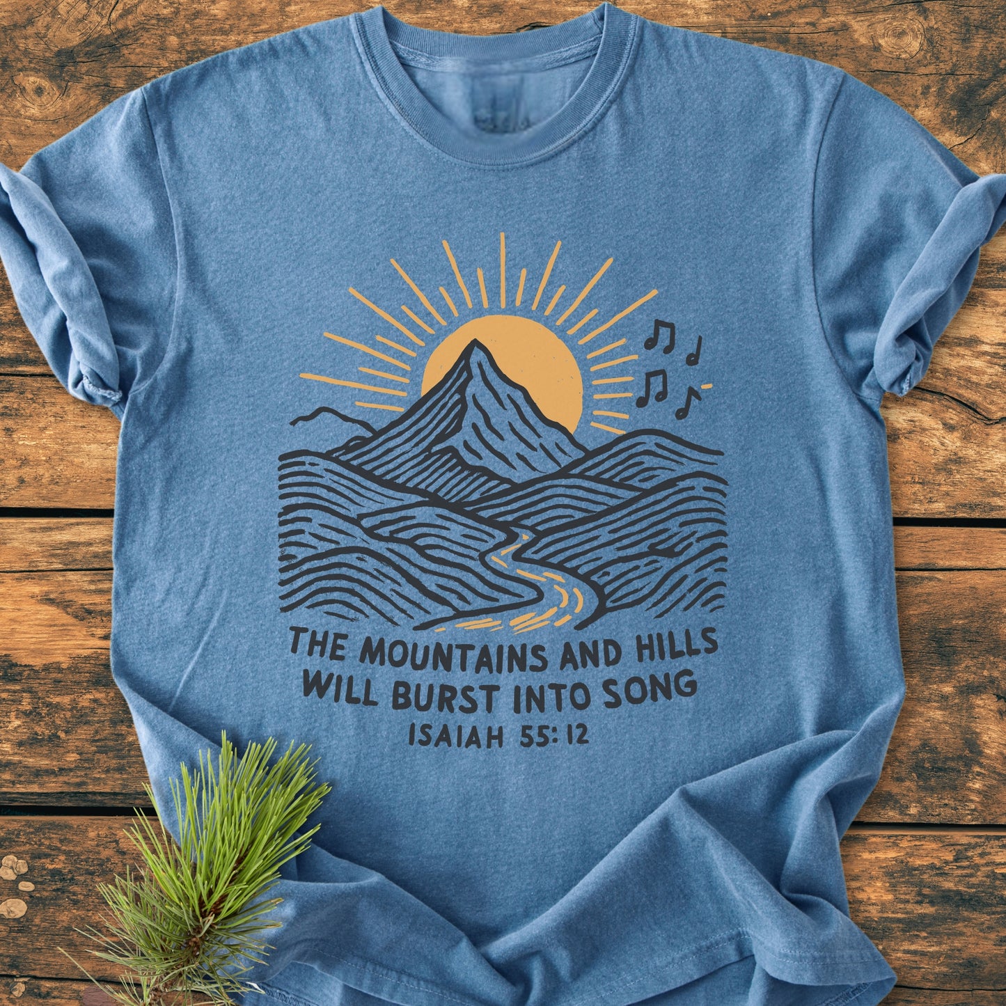 Mountains And Hills - Vintage Wash Tee features a stylized mountain graphic with musical notes and Isaiah 55:12 text, laid on wood beside a pine sprig, showcasing its relaxed fit.