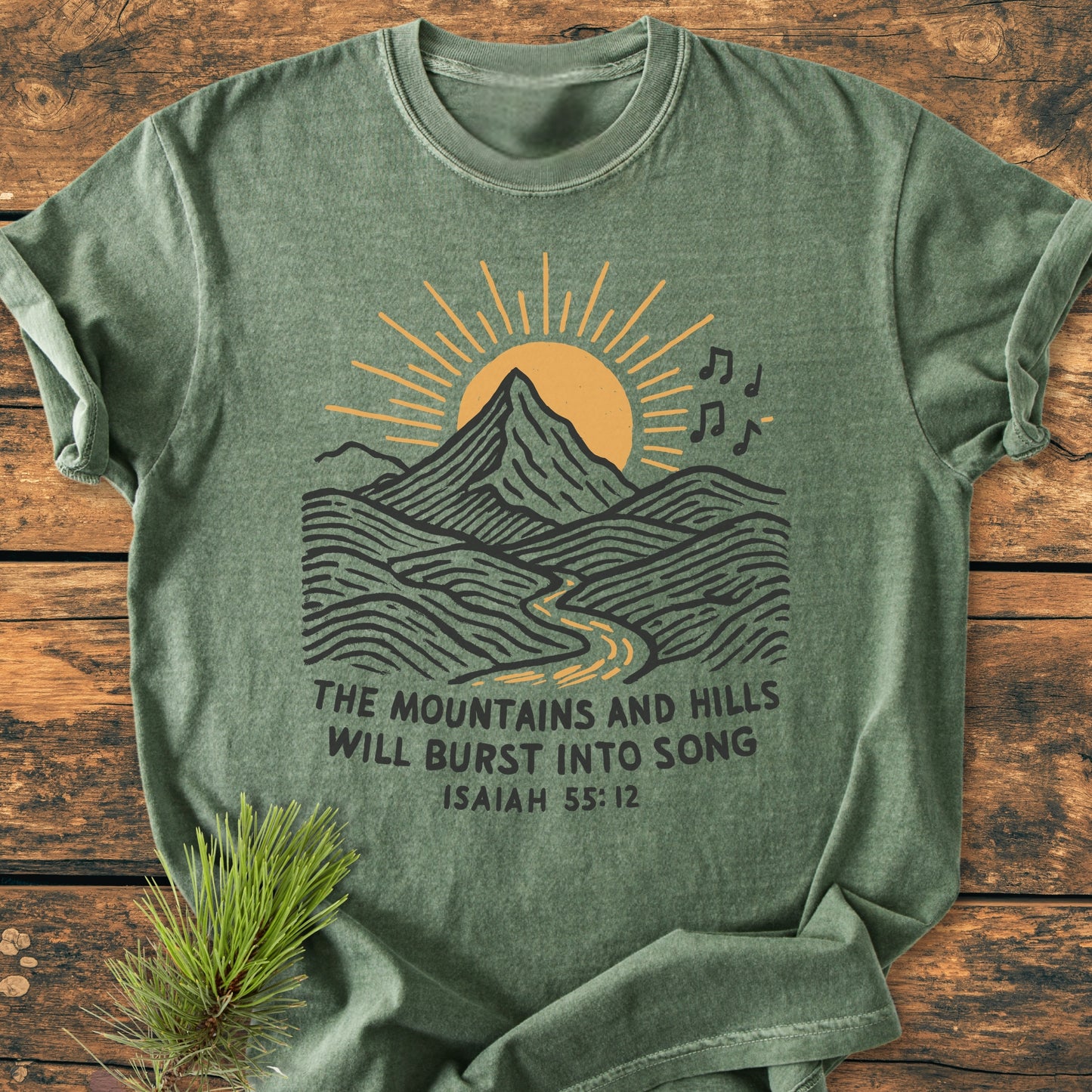 Mountains And Hills - Vintage Wash Tee: Green-heathered T-shirt featuring a mountain and sun design with musical notes, Isaiah 55:12 text, on a wooden surface, complemented by a pine sprig.