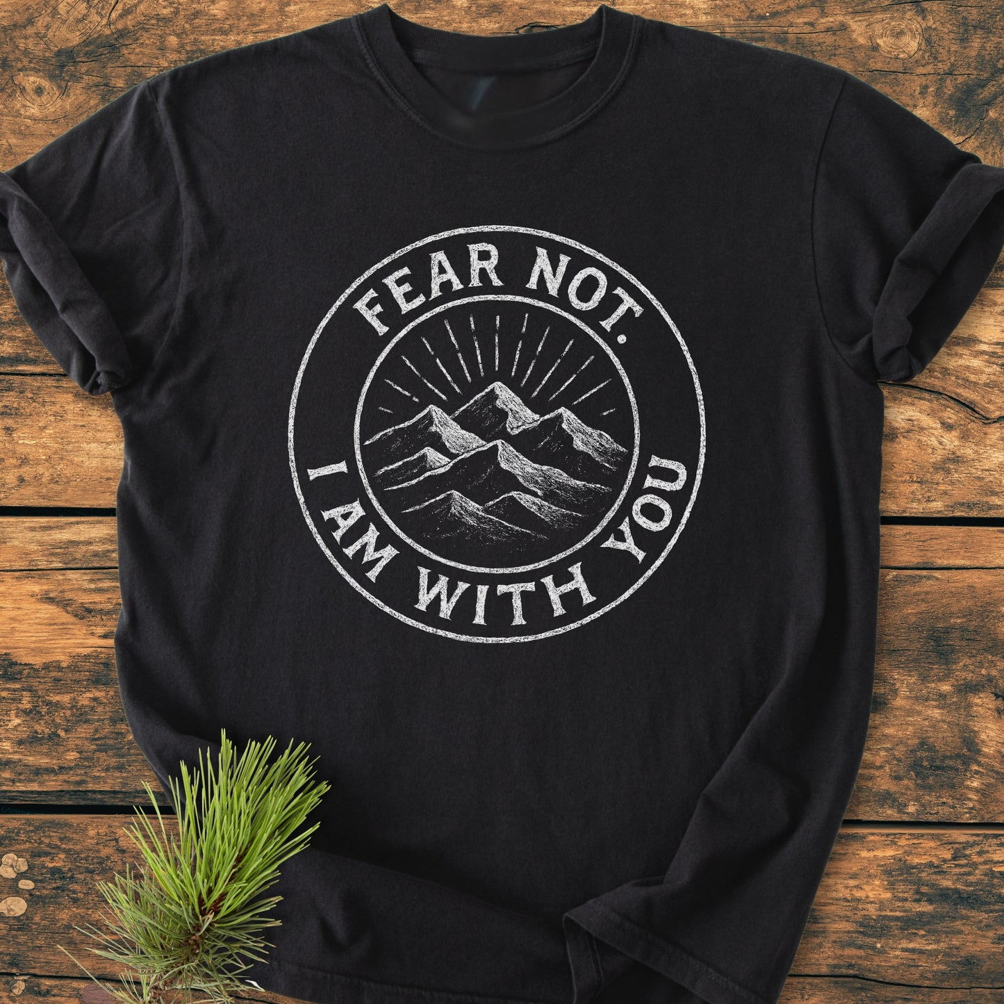 Fear Not - Vintage Wash Tee featuring a white chalk-style emblem of mountains and sun rays, laid on wood with a pine sprig beside, highlighting its rustic charm and cozy, relaxed fit.