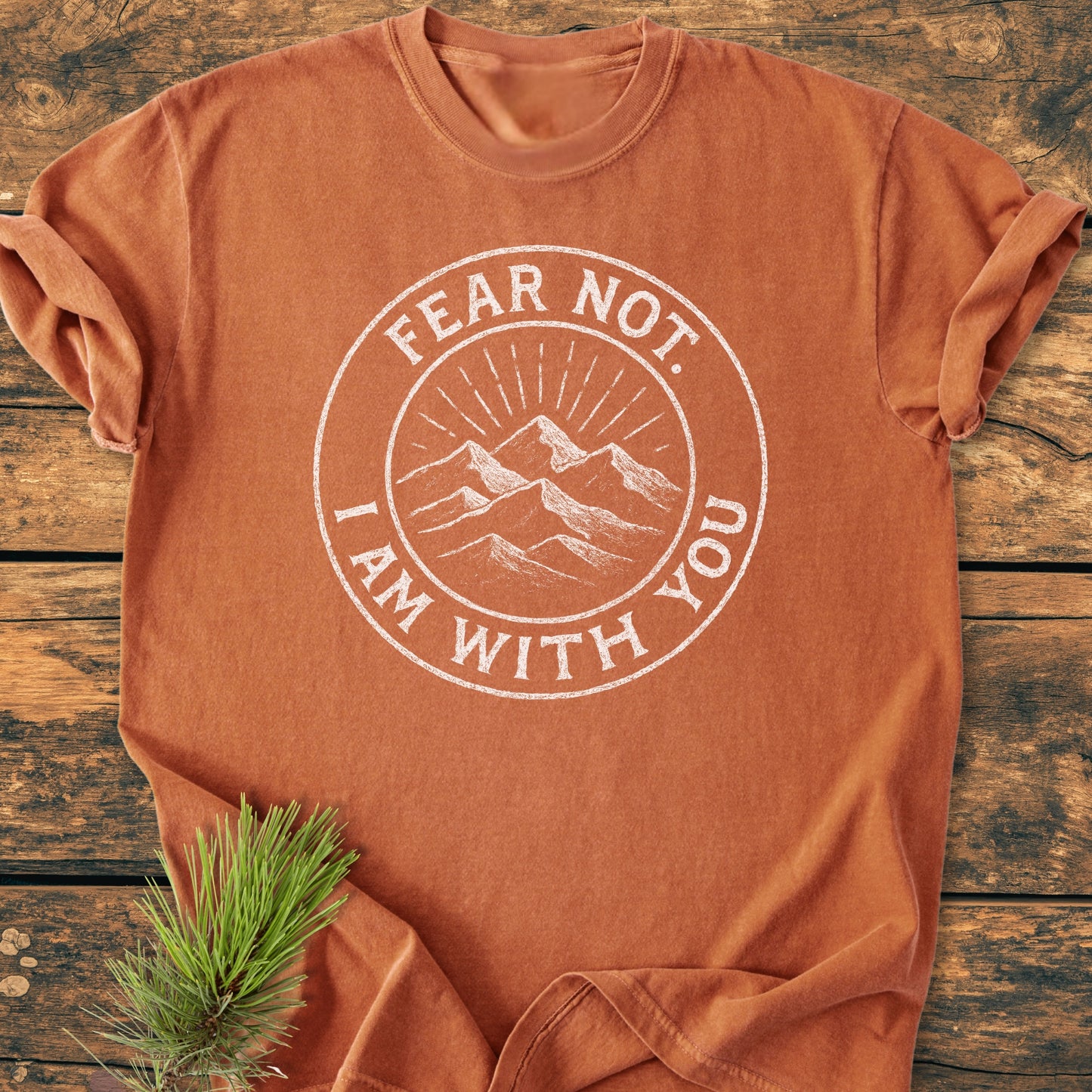 Fear Not - Vintage Wash Tee features a circular emblem with a mountain sketch and inspirational text. Laid flat on a wooden surface, it exudes comfort and durability with a relaxed fit.