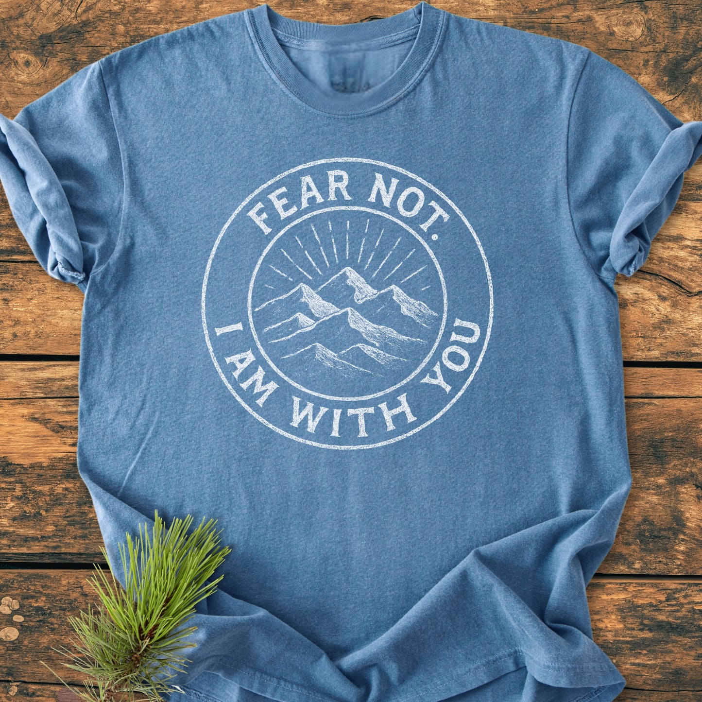 Heather-blue Fear Not - Vintage Wash Tee on wooden surface, featuring a white circular graphic of mountains and distressed lettering, FEAR NOT. I AM WITH YOU, with a pine branch accent.