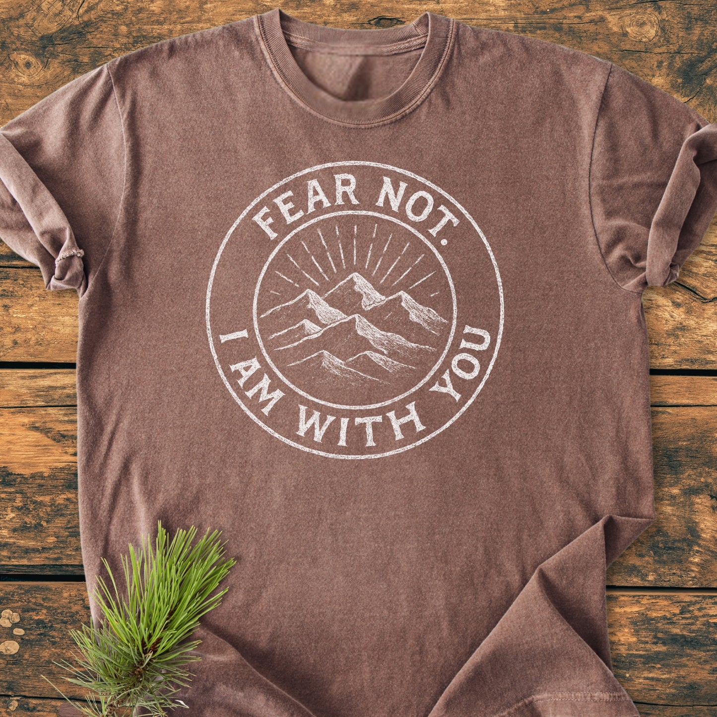 Fear Not - Vintage Wash Tee featuring white mountain graphic with text, Fear Not. I Am With You, laid flat on wood, accented by a pine sprig under the hem for a rustic touch.