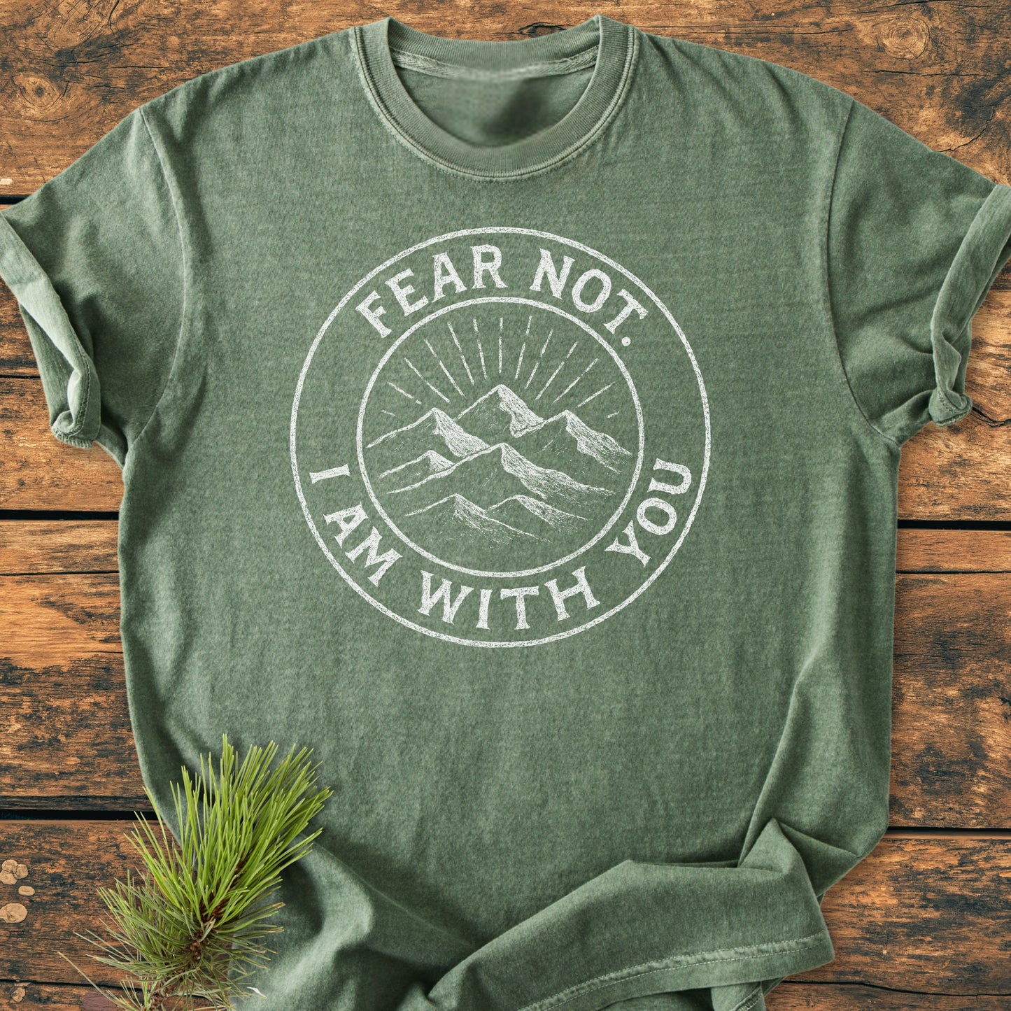 Fear Not - Vintage Wash Tee: A heathered green T-shirt with a white circular badge showing mountain peaks and the phrase “FEAR NOT. I AM WITH YOU,” laid on wood with a pine sprig accent.