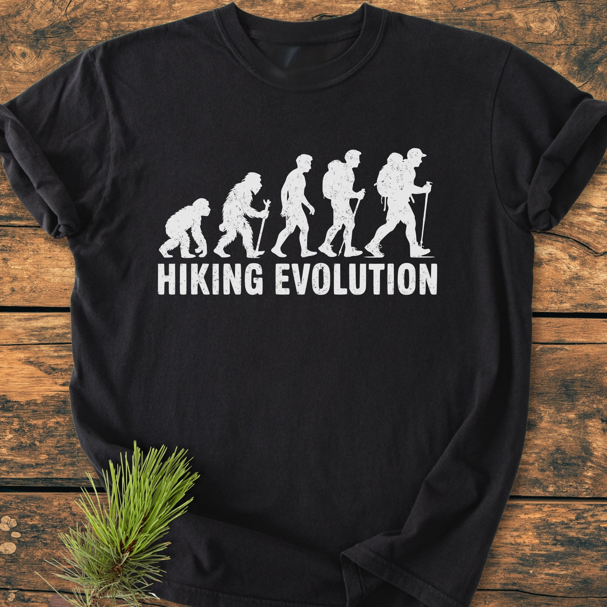 Hiking Evolution - Vintage Wash Tee featuring evolution of man graphic with hiking gear on a black T-shirt, laid on wood. Text HIKING EVOLUTION below. Pine branch accentuates natural theme.