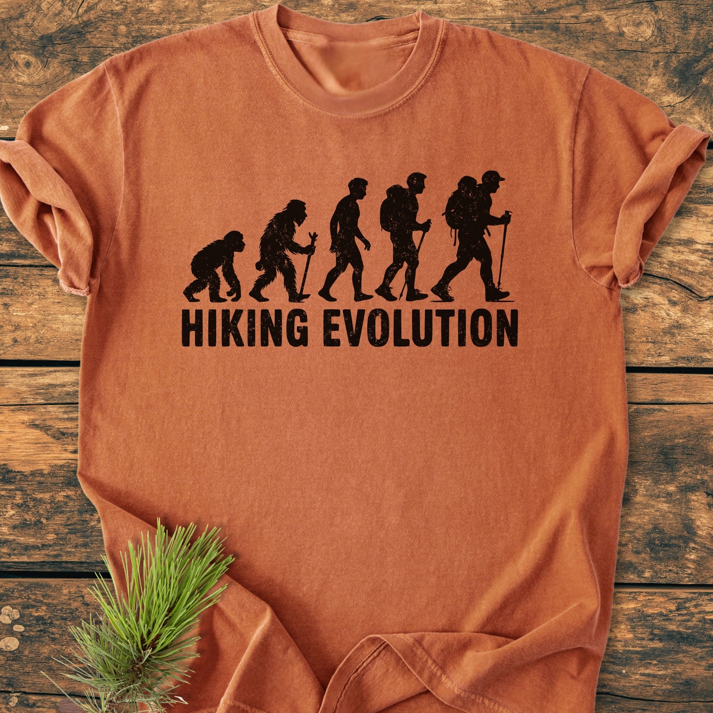 Hiking Evolution - Vintage Wash Tee: Burned-orange T-shirt with evolution of man hiking graphic and HIKING EVOLUTION text, lying flat on a wooden surface with a pine sprig for a rustic touch.