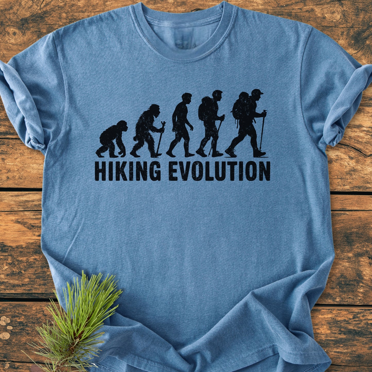 Hiking Evolution - Vintage Wash Tee: Light-blue t-shirt featuring ape-to-man evolution graphic ending with a hiker; text reads HIKING EVOLUTION. Displayed on wood with a pine branch accent.