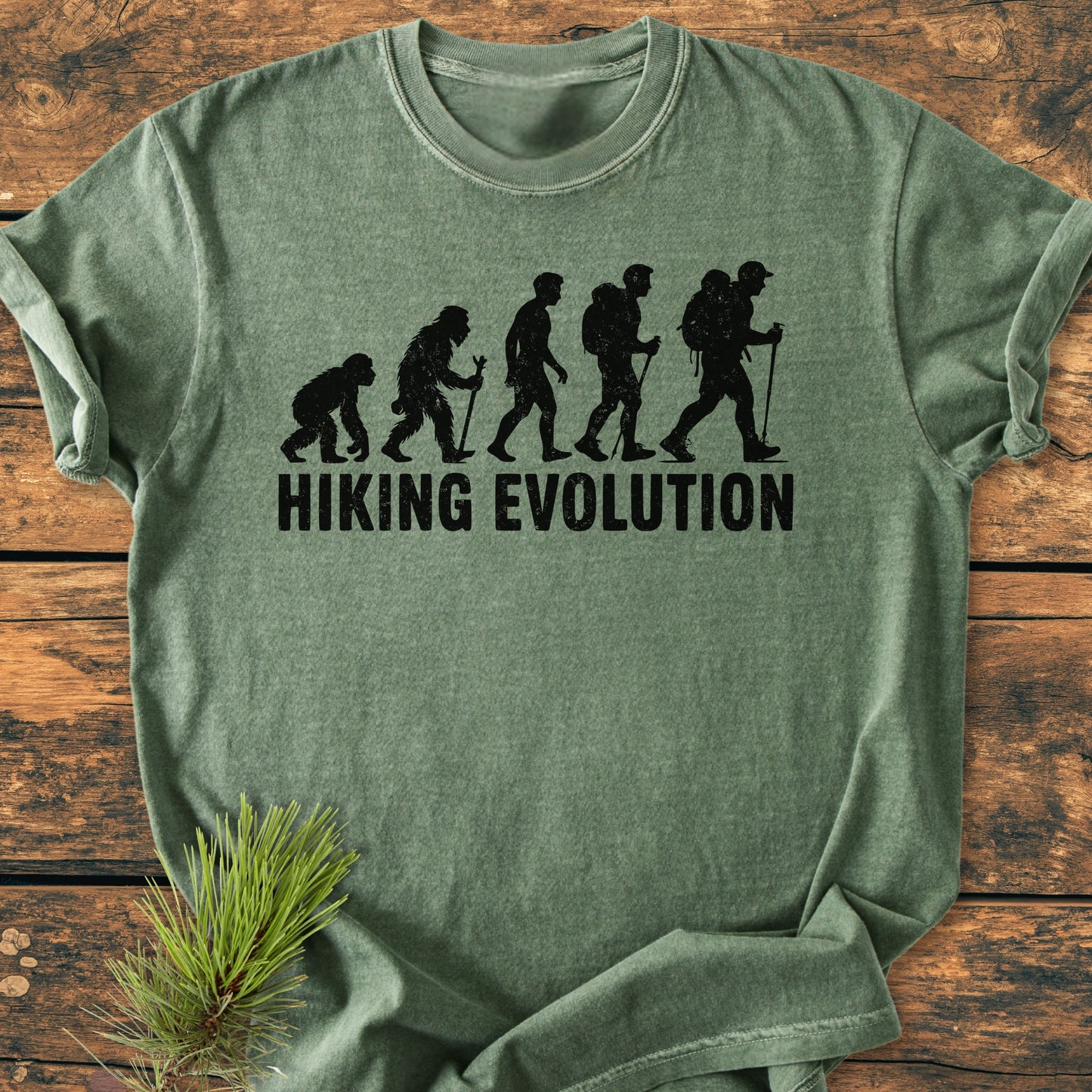 Hiking Evolution - Vintage Wash Tee features a graphic of the ape-to-modern human progression with a hiker, laid flat on wood, highlighting its outdoor theme with a pine sprig accent.