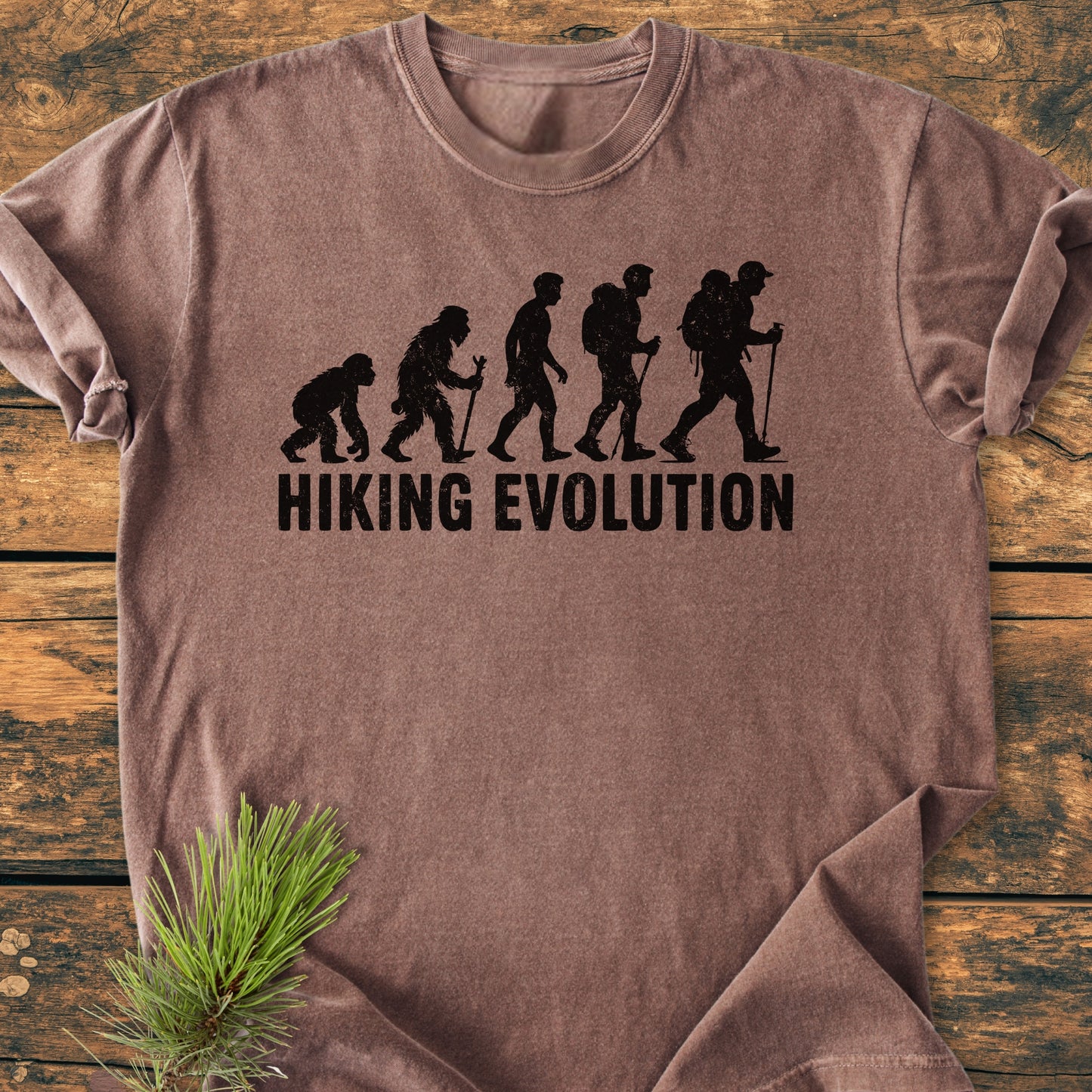 Hiking Evolution - Vintage Wash Tee features a black evolution of man hiking graphic on a brown-toned T-shirt, laid on wood with a pine branch accentuating its outdoorsy theme.
