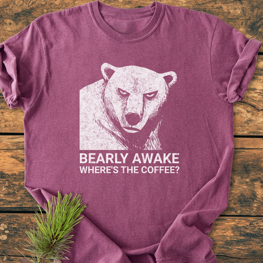 Bearly Awake - Vintage Wash Tee features a distressed bear graphic with BEARLY AWAKE WHERE’S THE COFFEE? caption, laid on a wooden surface with a pine sprig accent.