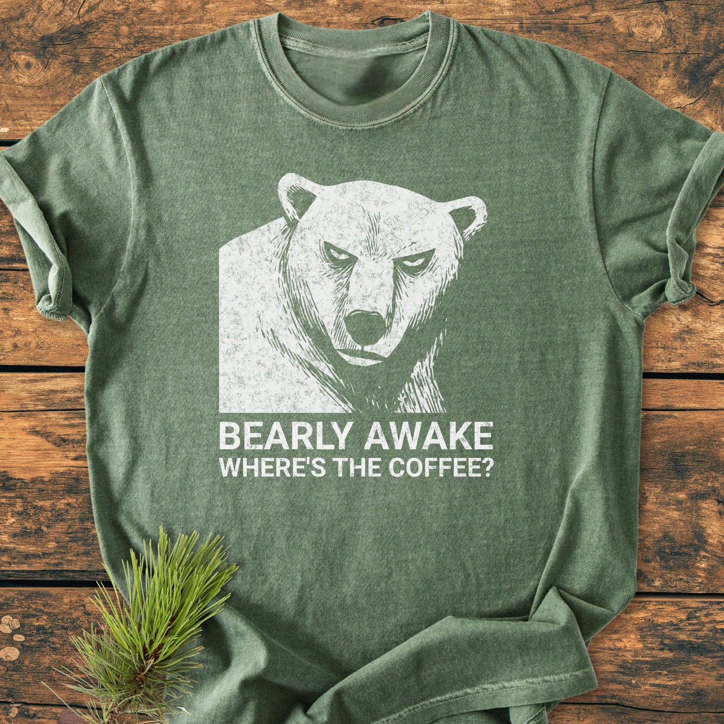 Bearly Awake - Vintage Wash Tee features a distressed bear graphic with the slogan “BEARLY AWAKE WHERE’S THE COFFEE?” on a forest-green shirt, set against a rustic wood surface with a pine sprig.
