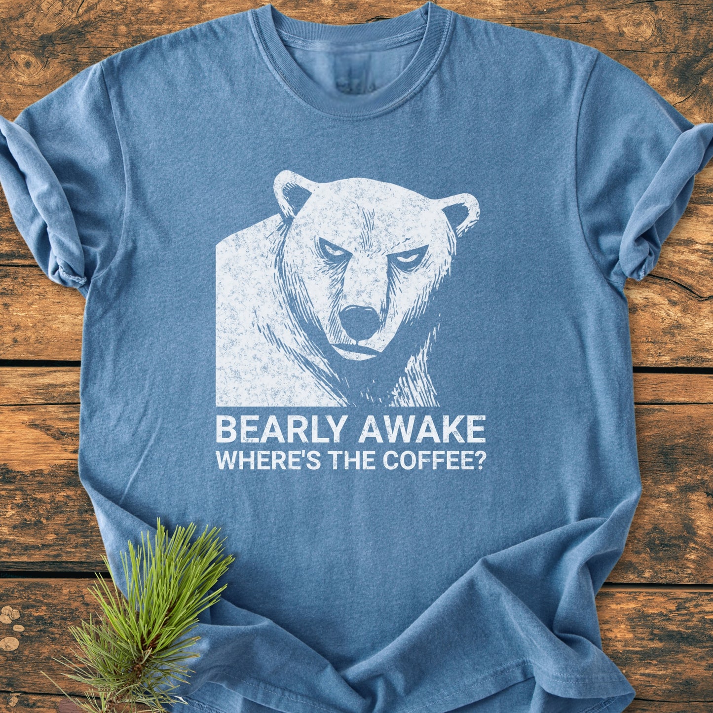 Bearly Awake - Vintage Wash Tee displays a distressed bear graphic and playful text on a heather-blue shirt, resting on wood with a pine sprig, highlighting its soft, garment-dyed cotton fabric.