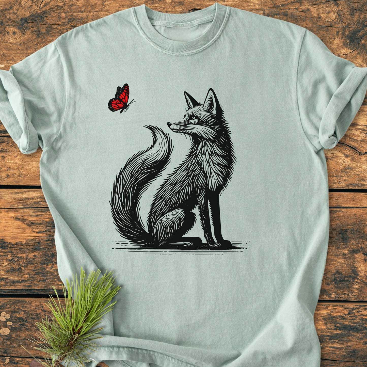 Fox And Butterfly - Vintage Wash Tee: Light-green crew-neck tee featuring a woodcut-style fox and butterfly illustration. Laid flat on rustic wood with a pine sprig beside it, showcasing its relaxed fit and durability.