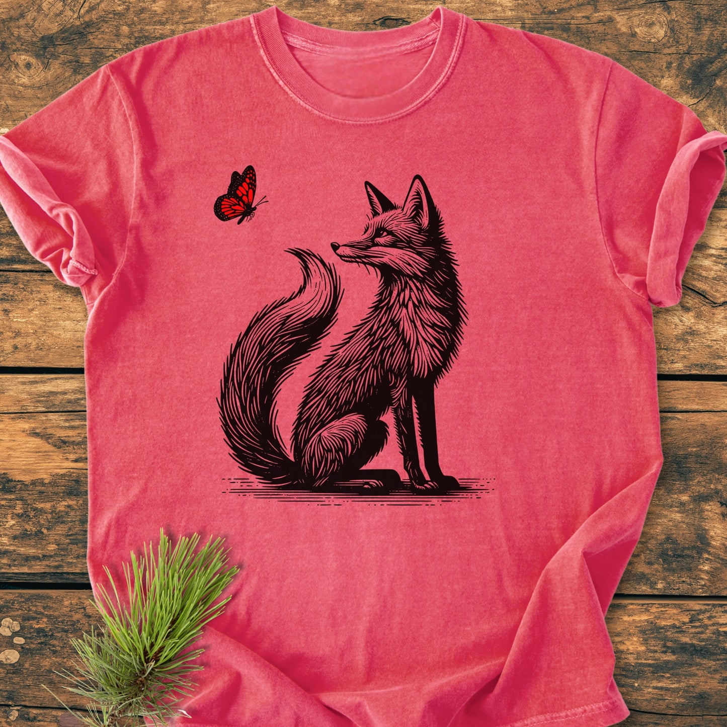 Fox And Butterfly - Vintage Wash Tee: A heathered red T-shirt featuring a black-ink illustration of a fox gazing at a red butterfly, displayed on a rustic wooden surface with a pine sprig accent.