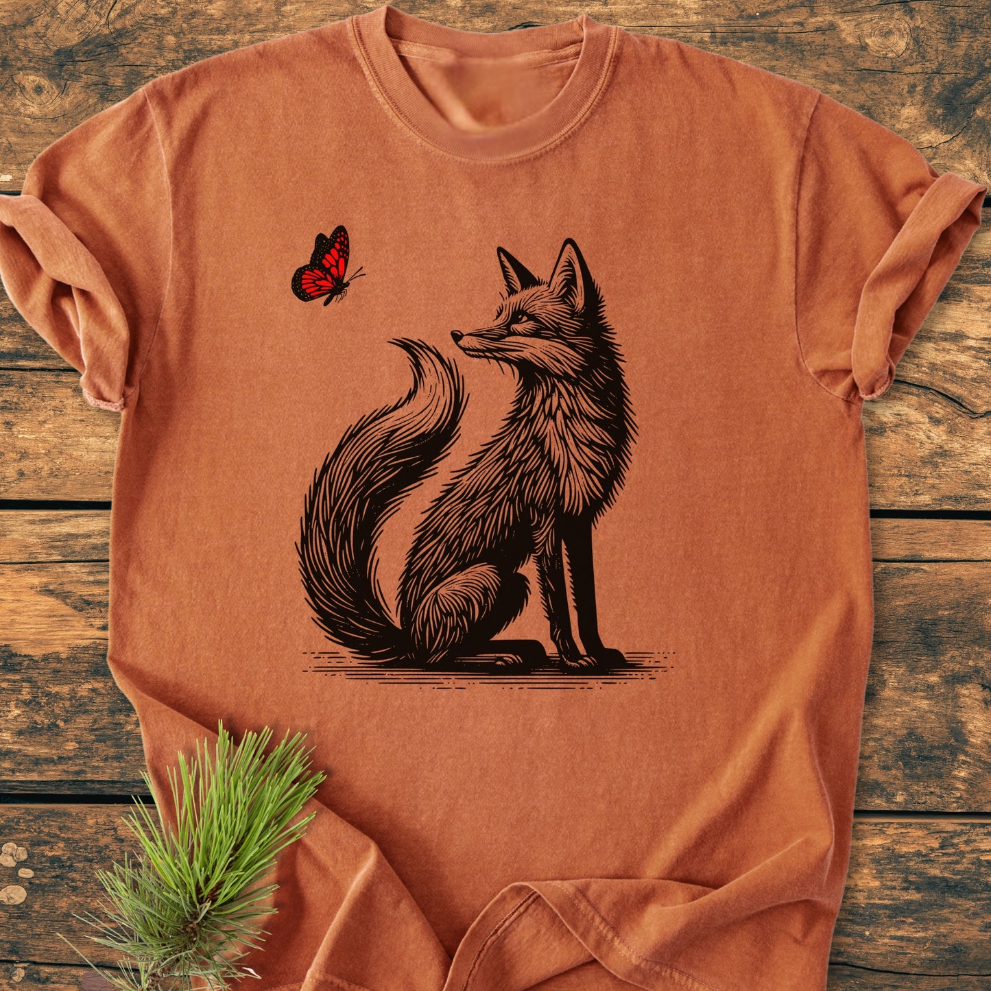 Fox And Butterfly - Vintage Wash Tee features a stylized fox and butterfly print on a rust-orange T-shirt, laid on a wooden surface with a pine twig beside it.