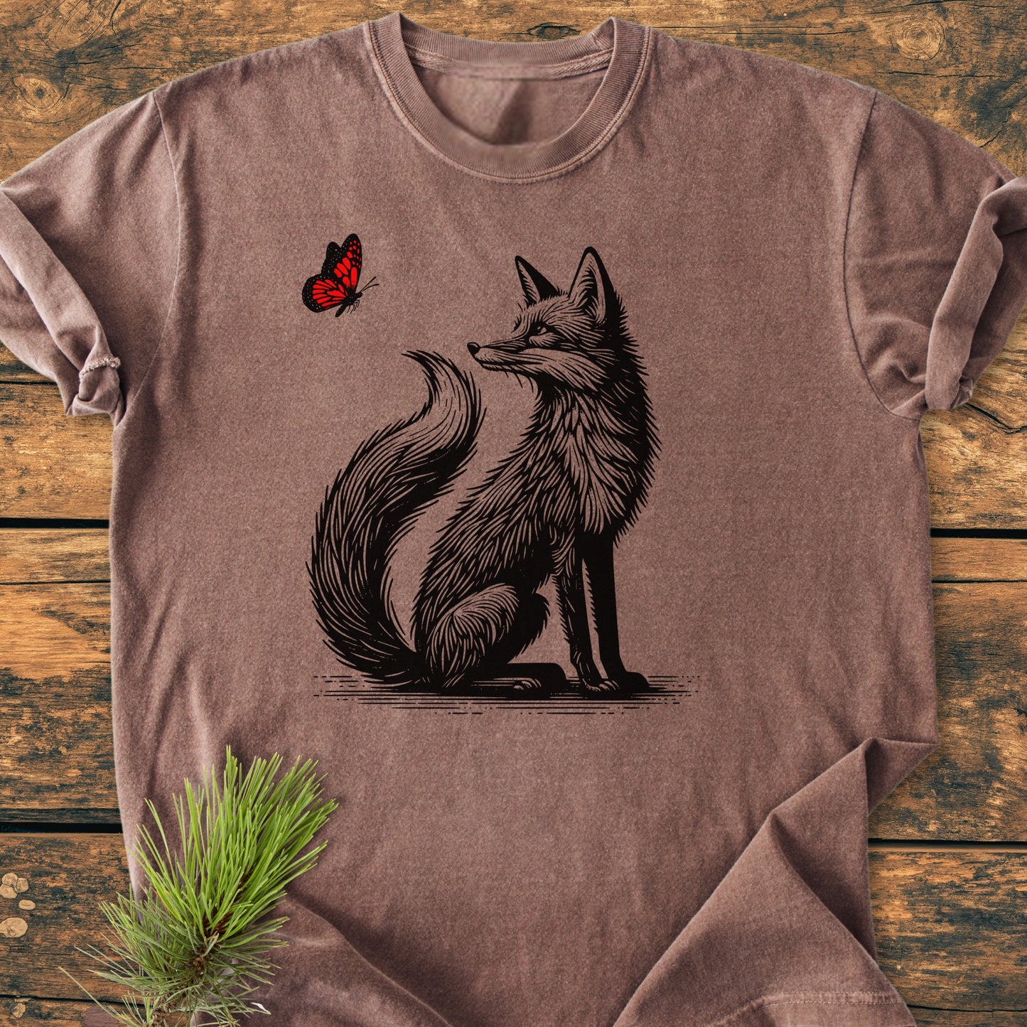 Fox And Butterfly - Vintage Wash Tee, featuring a black line-art fox gazing at a red butterfly, displayed on a brown short-sleeve shirt, lying flat on a wooden surface with a pine branch nearby.