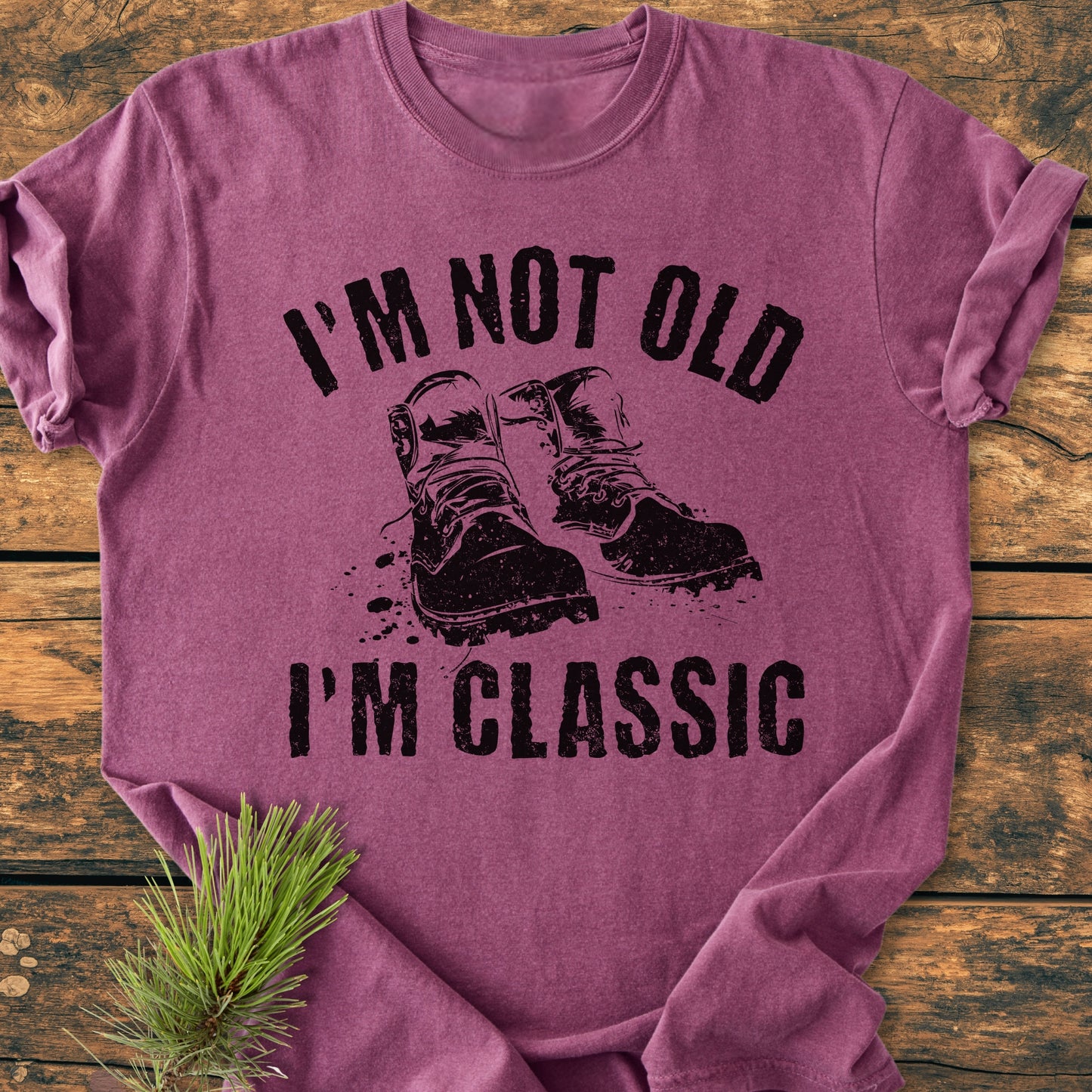 I'm Not Old I'm Classic - Vintage Wash Tee, a heather-purple crew-neck shirt with bold black lettering and a rugged boot graphic, laid on a wooden surface alongside a pine sprig.