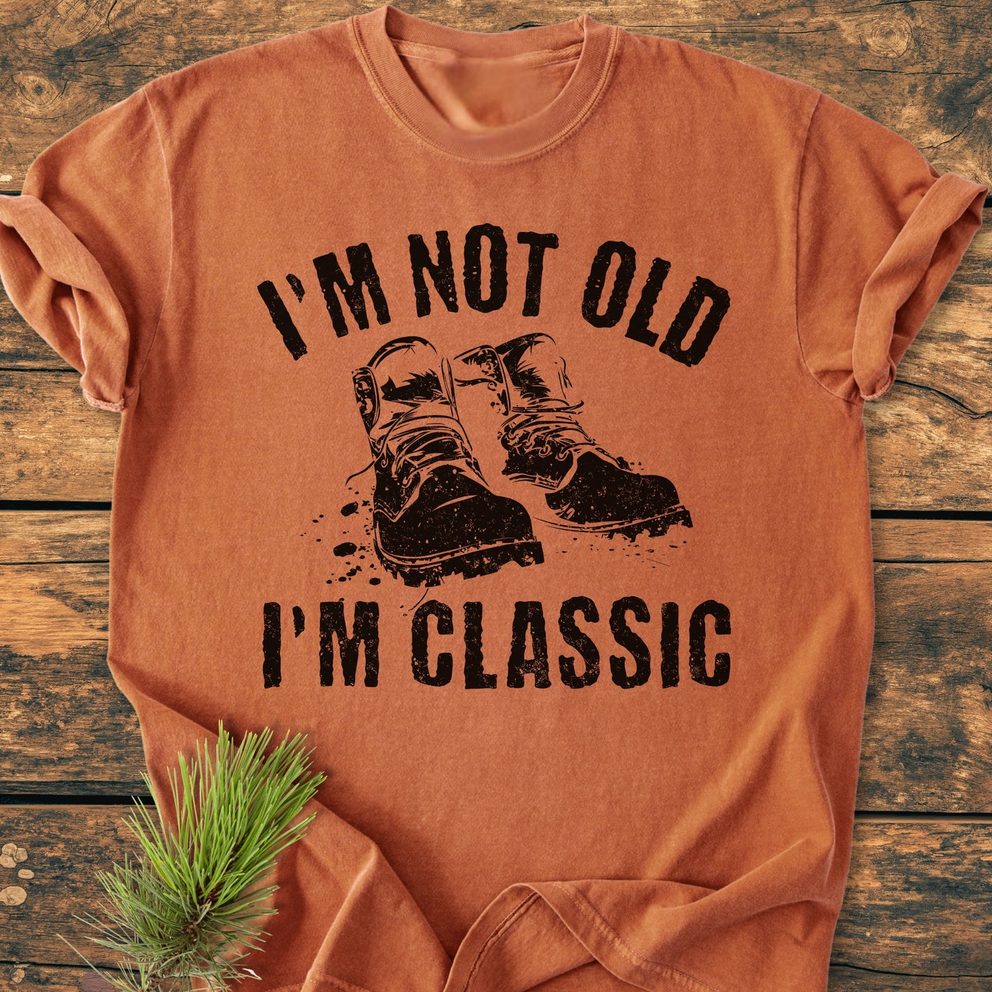 I'm Not Old I'm Classic - Vintage Wash Tee featuring bold black lettering and work boots graphic on soft, relaxed-fit Comfort Colors garment-dyed fabric, displayed with an evergreen sprig on wooden surface.