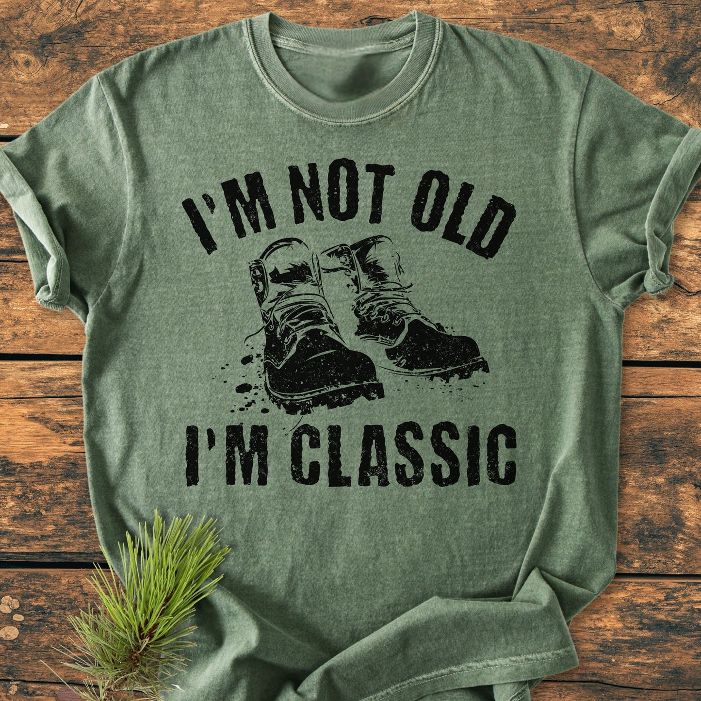 Heather-green I'm Not Old I'm Classic - Vintage Wash Tee featuring a black boots graphic and slogan, laid on a wooden surface with a pine sprig beside it.