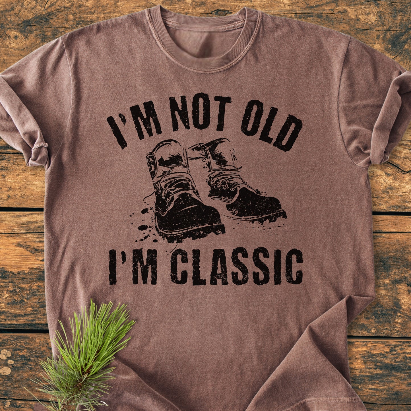 I'm Not Old I'm Classic - Vintage Wash Tee featuring a distressed work boots graphic, displayed on a rustic wooden surface with a pine sprig accent. Short-sleeve, crewneck, relaxed fit, durable stitching.