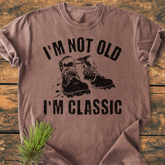 I'm Not Old I'm Classic - Vintage Wash Tee featuring a distressed work boots graphic, displayed on a rustic wooden surface with a pine sprig accent. Short-sleeve, crewneck, relaxed fit, durable stitching.