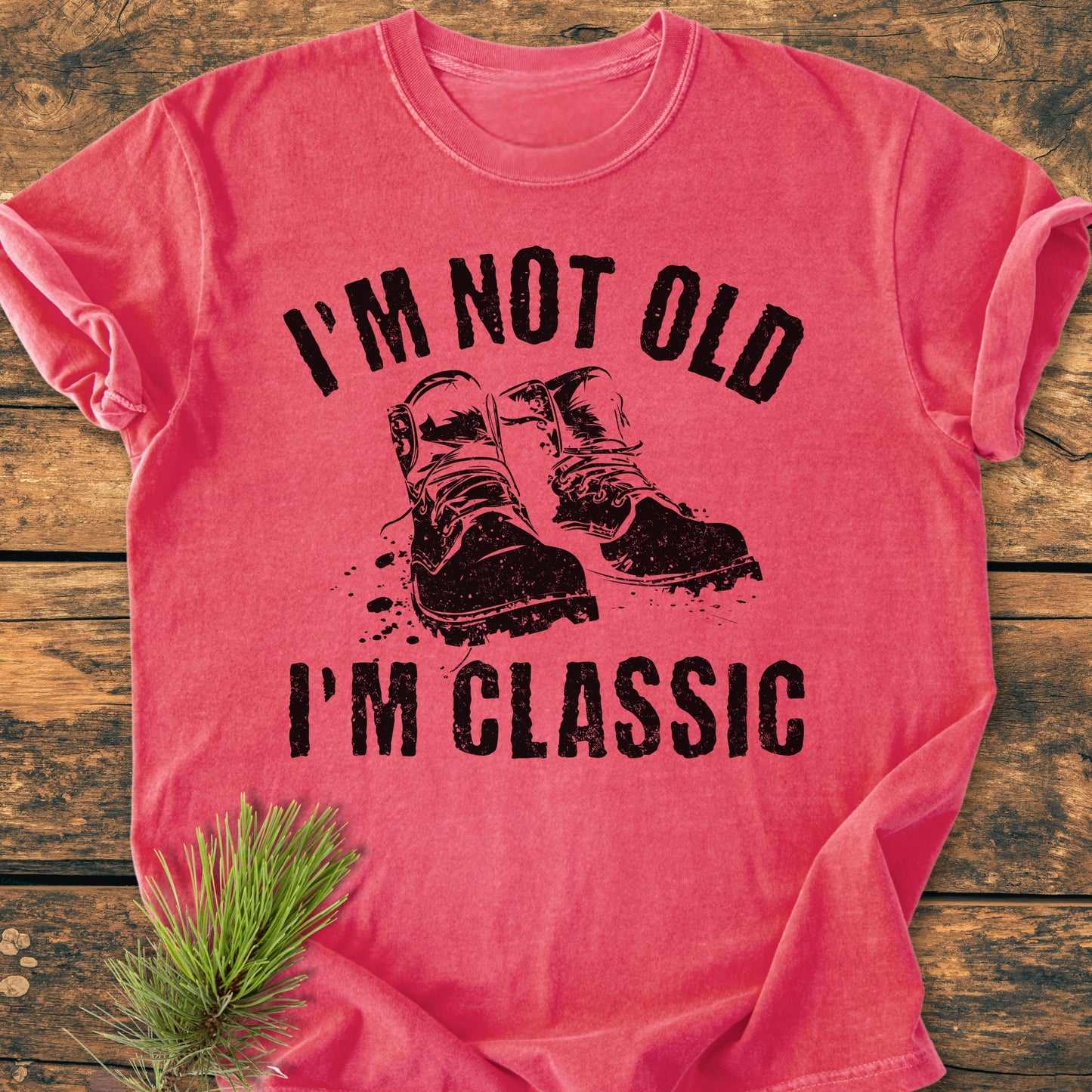 I'm Not Old I'm Classic - Vintage Wash Tee in heathered red/pink, featuring rugged boots graphic and slogan, laid on wooden surface with pine sprig, showcasing the tee's relaxed fit and soft-washed texture.