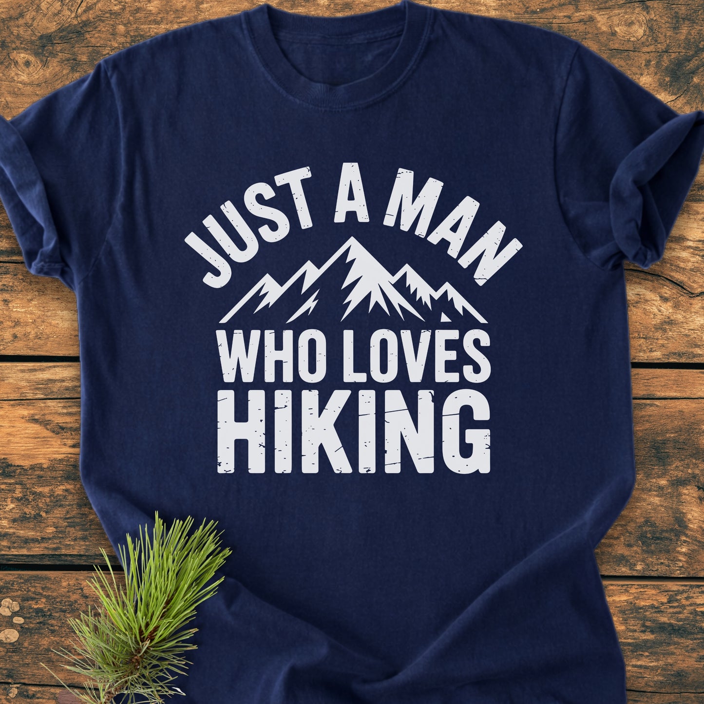 Navy-blue Just A Man Who Loves Hiking - Vintage Wash Tee featuring distressed white mountain graphics and slogan, laid flat on wood with a pine sprig, showcasing its relaxed fit and durable stitching.
