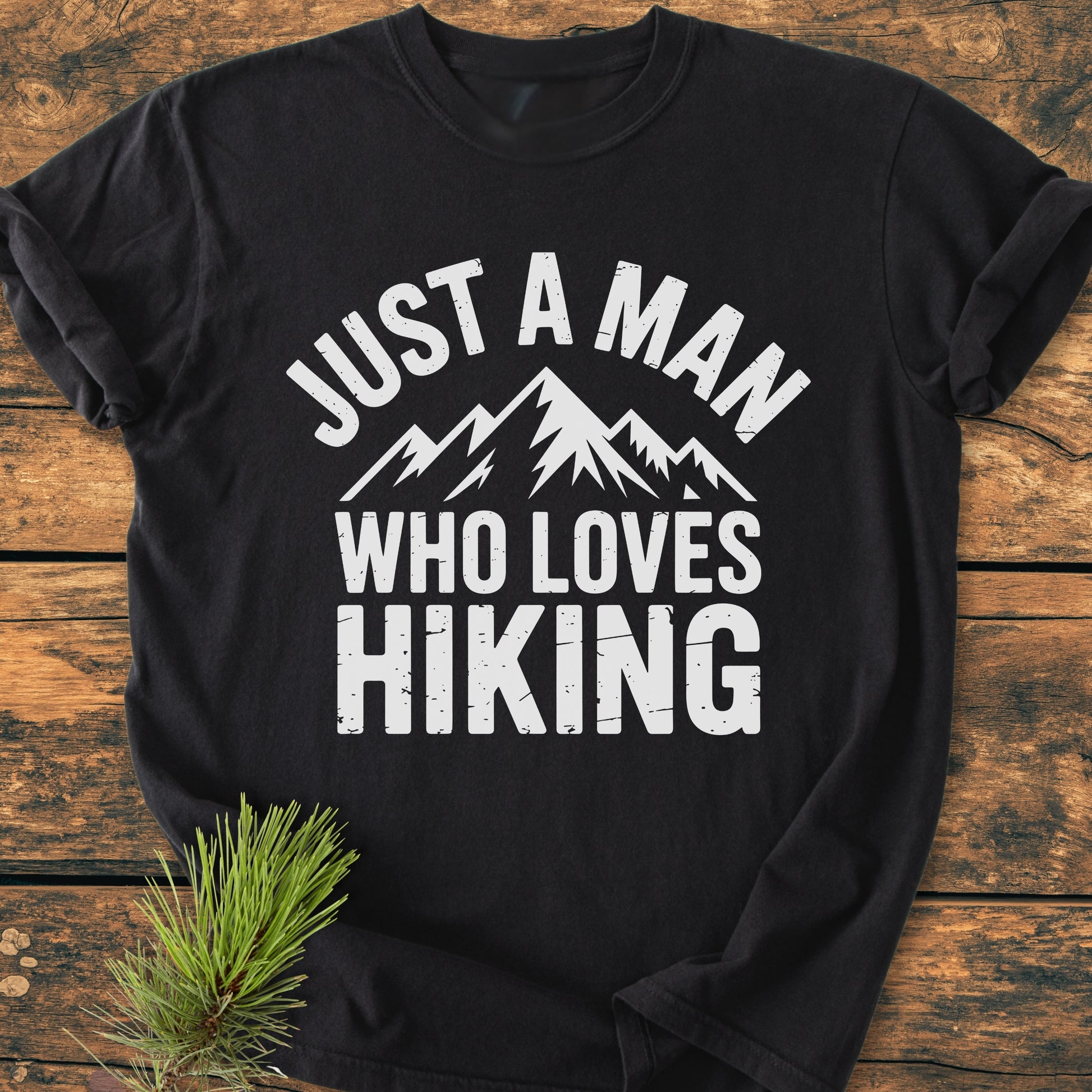 Just A Man Who Loves Hiking - Vintage Wash Tee displays stylized mountain peaks and slogan on a black T-shirt, laid on rustic wood with a pine sprig beside, highlighting its cozy, durable design.