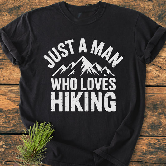 Just A Man Who Loves Hiking - Vintage Wash Tee displays stylized mountain peaks and slogan on a black T-shirt, laid on rustic wood with a pine sprig beside, highlighting its cozy, durable design.