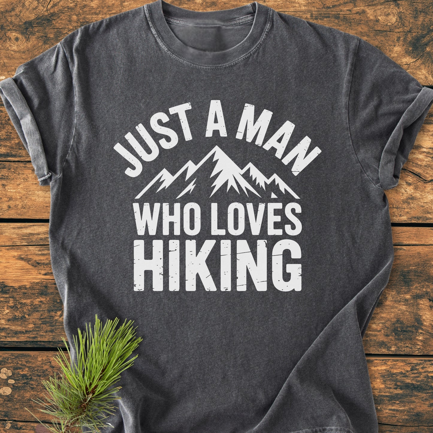 Just A Man Who Loves Hiking - Vintage Wash Tee featuring a white stylized mountain graphic. Laid on a wooden surface, accompanied by a pine twig, emphasizing the outdoor theme.