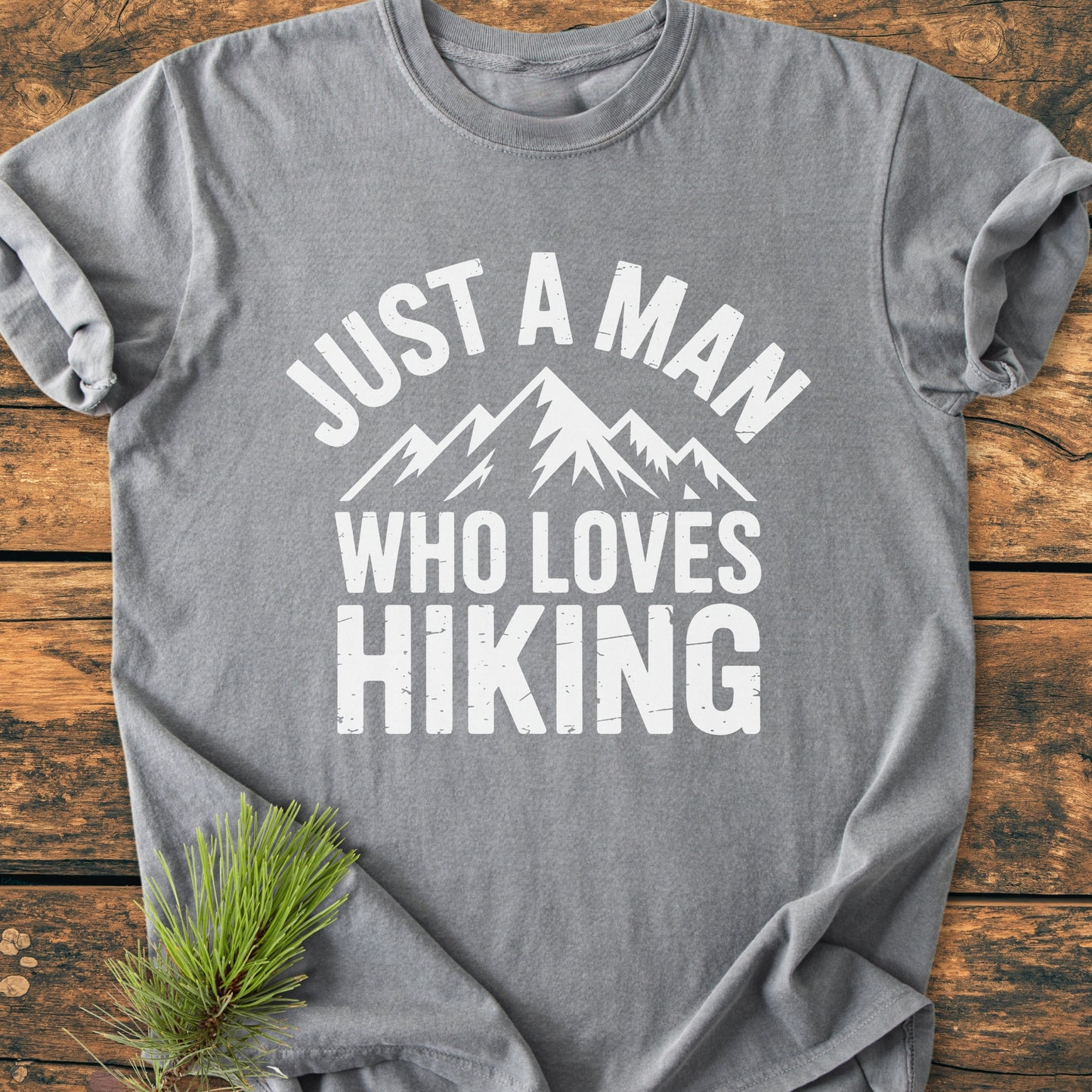 Heather-gray Just A Man Who Loves Hiking - Vintage Wash Tee with bold white lettering and mountain graphic, laid flat on wood. Pine sprig accentuates outdoor theme. Made from soft, durable ring-spun cotton.