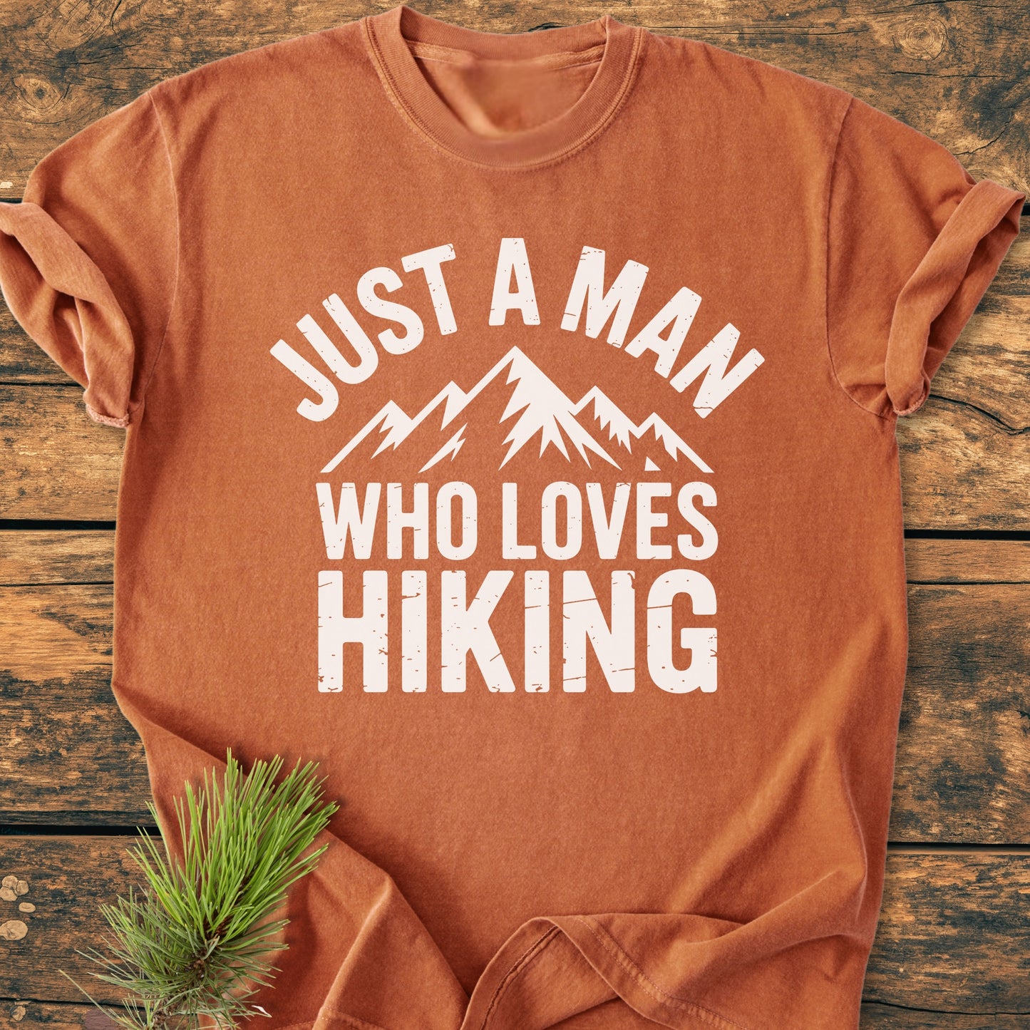 Burnt-orange Just A Man Who Loves Hiking - Vintage Wash Tee folded on wood, featuring white lettering and mountain graphic, accompanied by a pine sprig, showcasing its soft, relaxed fit and durability.