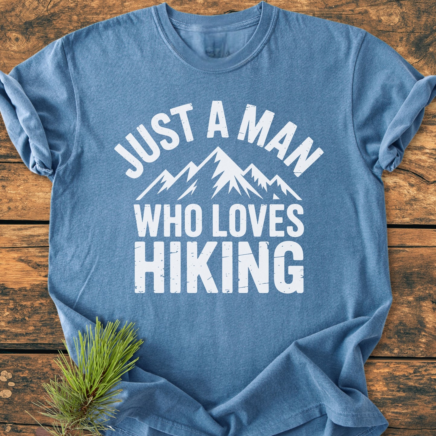 Light-blue Just A Man Who Loves Hiking T-shirt featuring mountain graphic, laid on wood. Includes pine sprig accent. Made from soft, durable, pre-shrunk 100% ring-spun cotton for comfort.