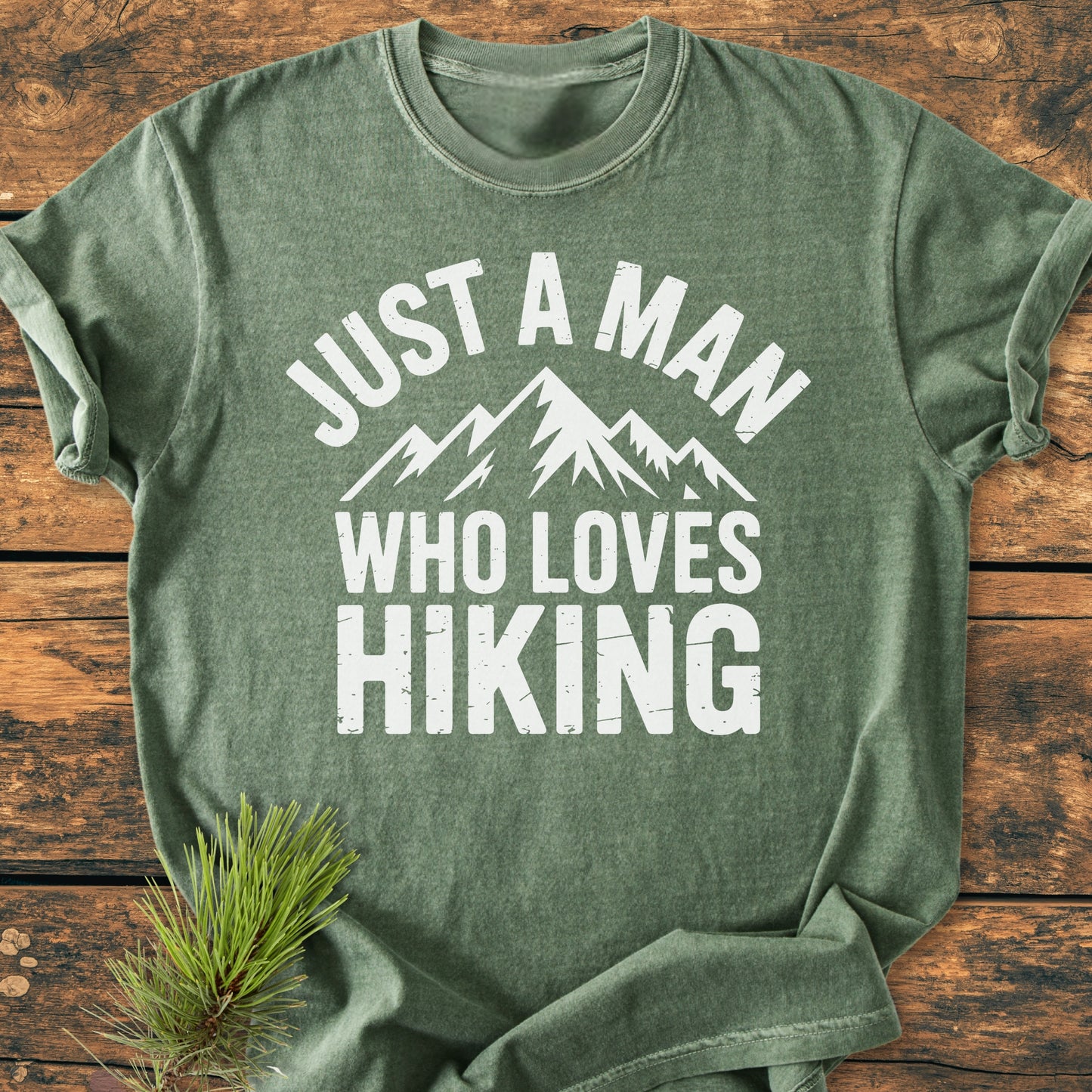 Just A Man Who Loves Hiking - Vintage Wash Tee: Heather-green T-shirt with bold white lettering and mountain graphic, accented by pine needles on a rustic wooden surface, showcasing its outdoorsy theme and relaxed fit.