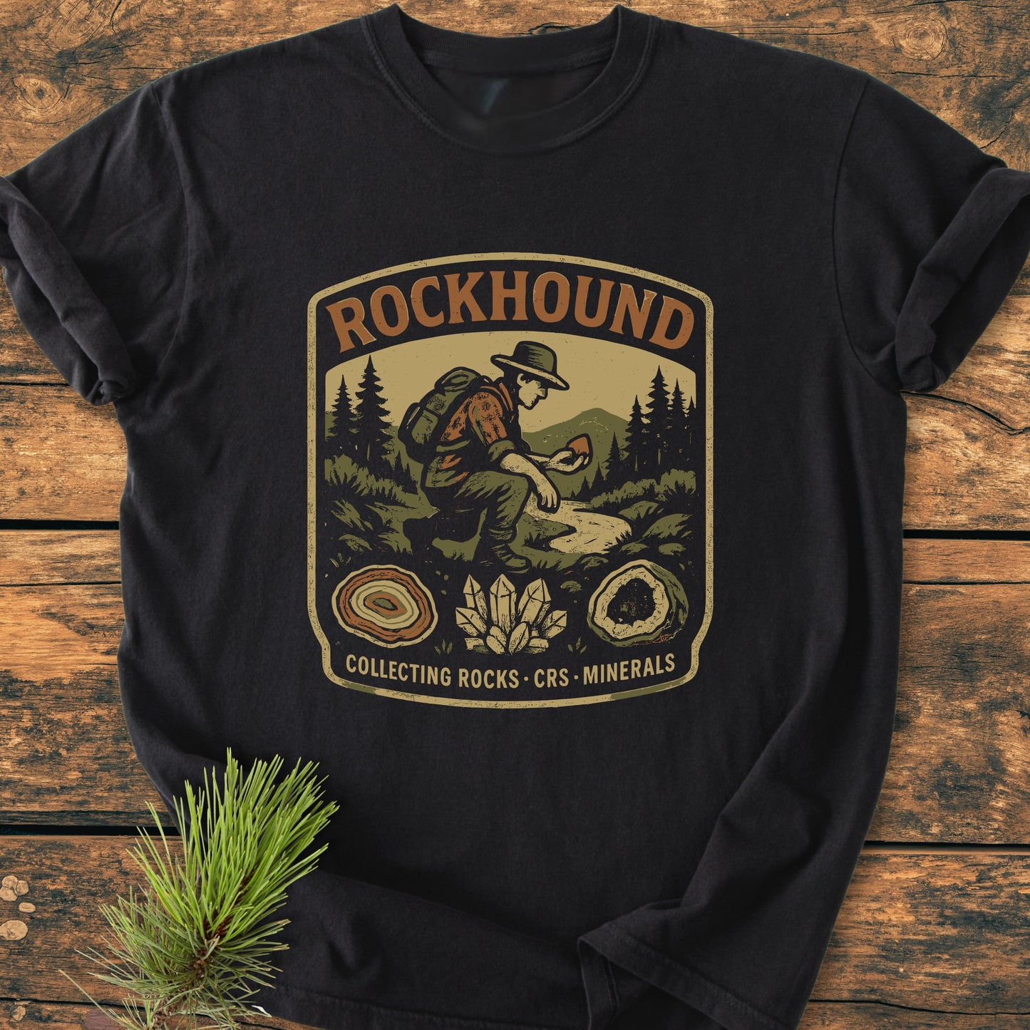 Rockhound Badge - Vintage Wash Tee featuring a hiker graphic with evergreen trees, mountains, and rock illustrations on a black T-shirt, laid on a wooden surface. Comfortable, durable, garment-dyed cotton.