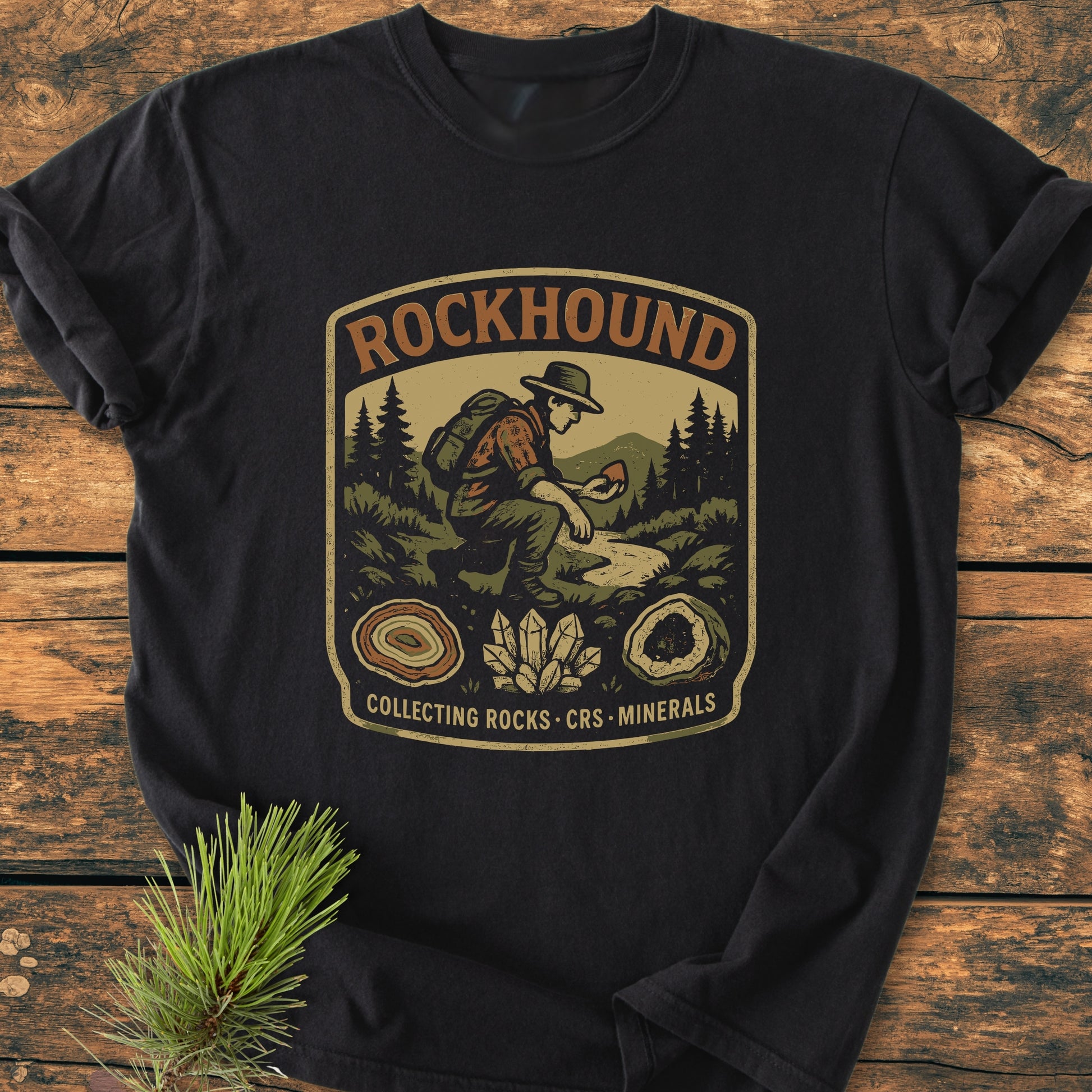 Rockhound Badge - Vintage Wash Tee featuring a hiker graphic with evergreen trees, mountains, and rock illustrations on a black T-shirt, laid on a wooden surface. Comfortable, durable, garment-dyed cotton.