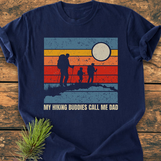 My Hiking Buddies Call Me Dad - Vintage Wash Tee features a retro sunset graphic with silhouettes of hikers, laid on a wooden surface, highlighting its soft, durable 100% ring-spun cotton construction.