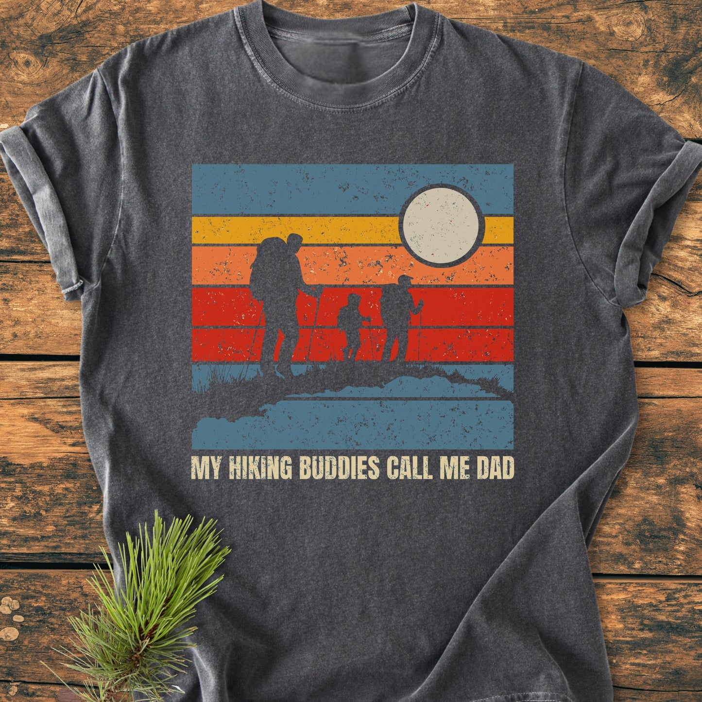 Charcoal-gray My Hiking Buddies Call Me Dad - Vintage Wash Tee featuring a retro graphic of hikers and sun. Displayed on wood surface with pine sprig. Cozy, relaxed fit, perfect for hiking enthusiasts.
