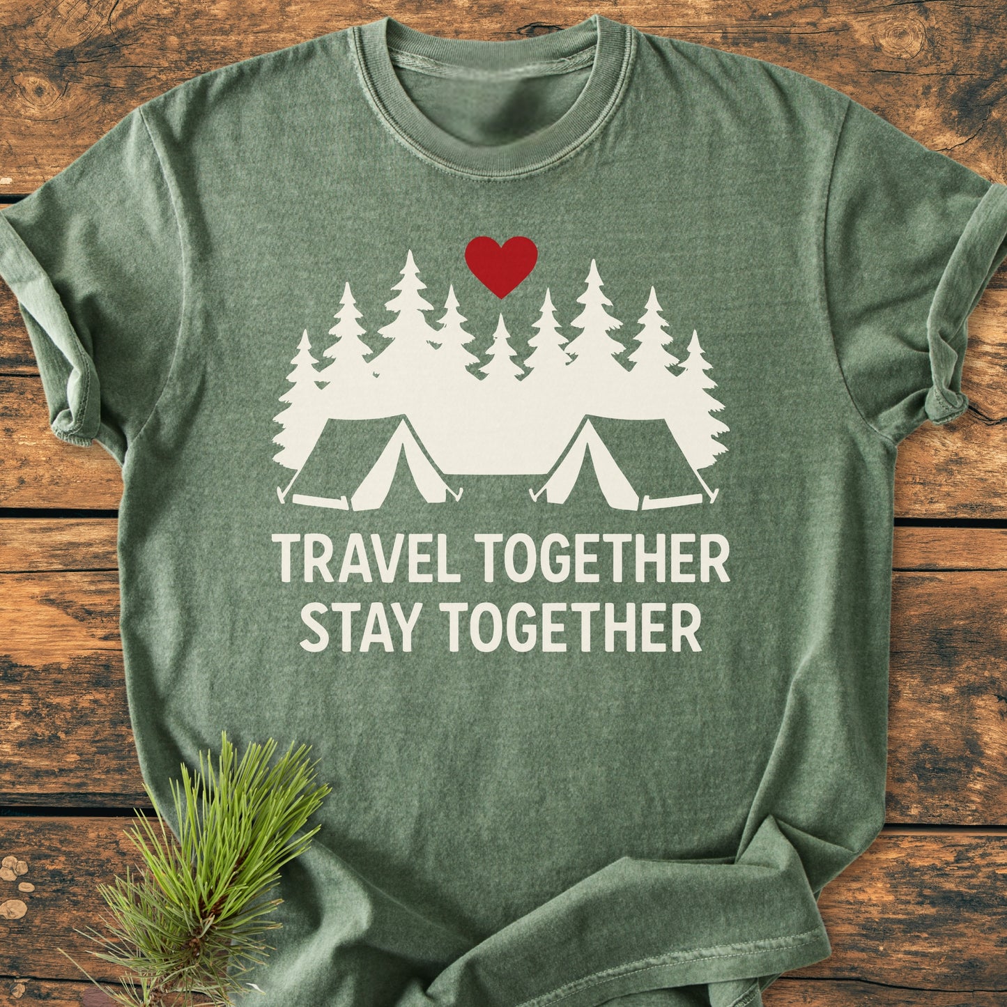Travel Together Stay Together - Vintage Wash Tee features a white silhouette of pine trees and tents with a red heart on a green T-shirt, laid on wood with a pine sprig.