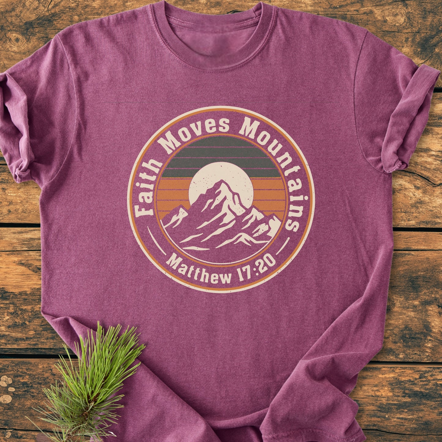 Faith Moves Mountains - Vintage Wash Tee in heather-purple, featuring a mountain graphic with inspirational text. Laid flat on rustic wood, highlighting its relaxed fit and durable double-needle stitching.