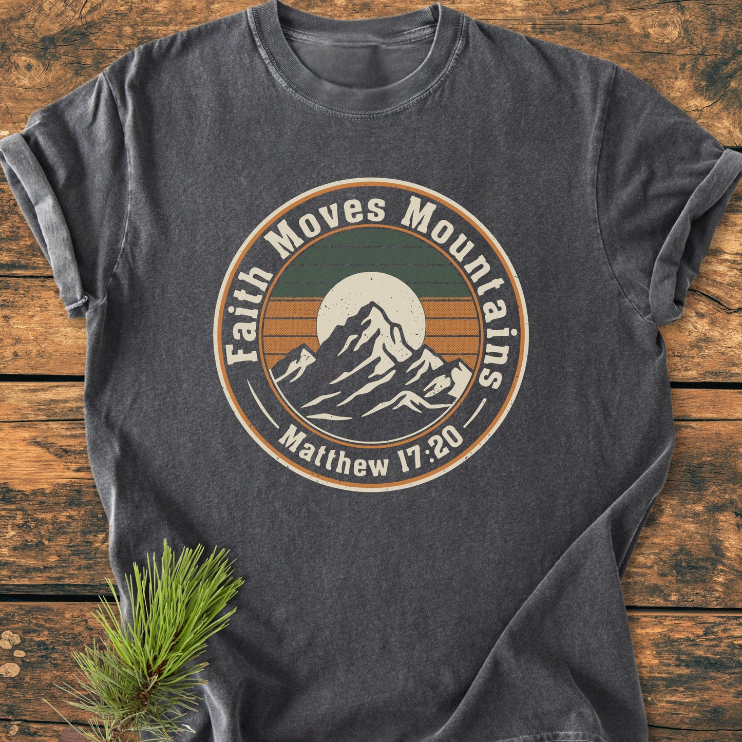 Faith Moves Mountains - Vintage Wash Tee featuring a circular emblem of snow-capped mountains with a sun and stripes, surrounded by Matthew 17:20 text, laid on a rustic wooden surface.