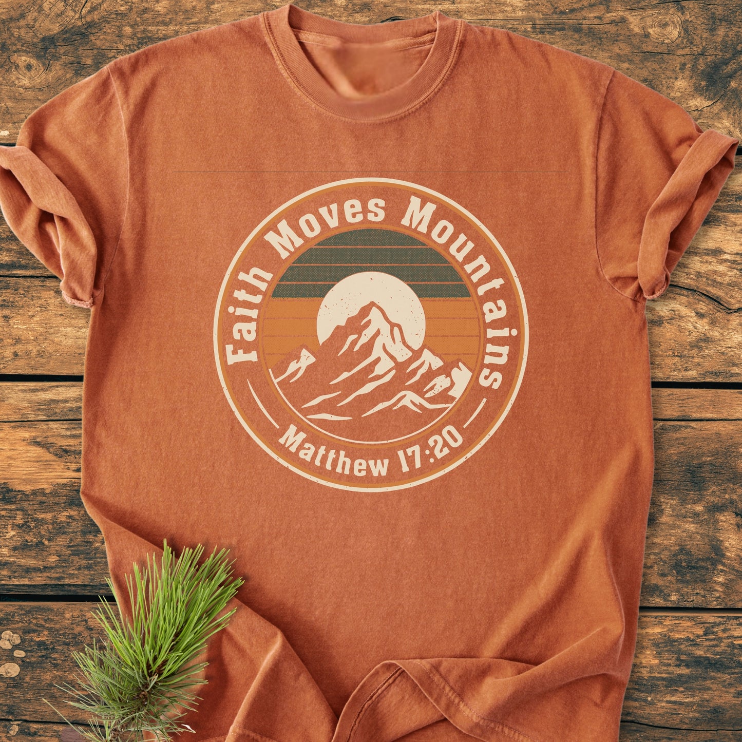 Faith Moves Mountains - Vintage Wash Tee, rust-orange with a sunset mountain graphic and Bible reference. Laid flat with an evergreen sprig, highlighting its rustic, outdoorsy appeal.