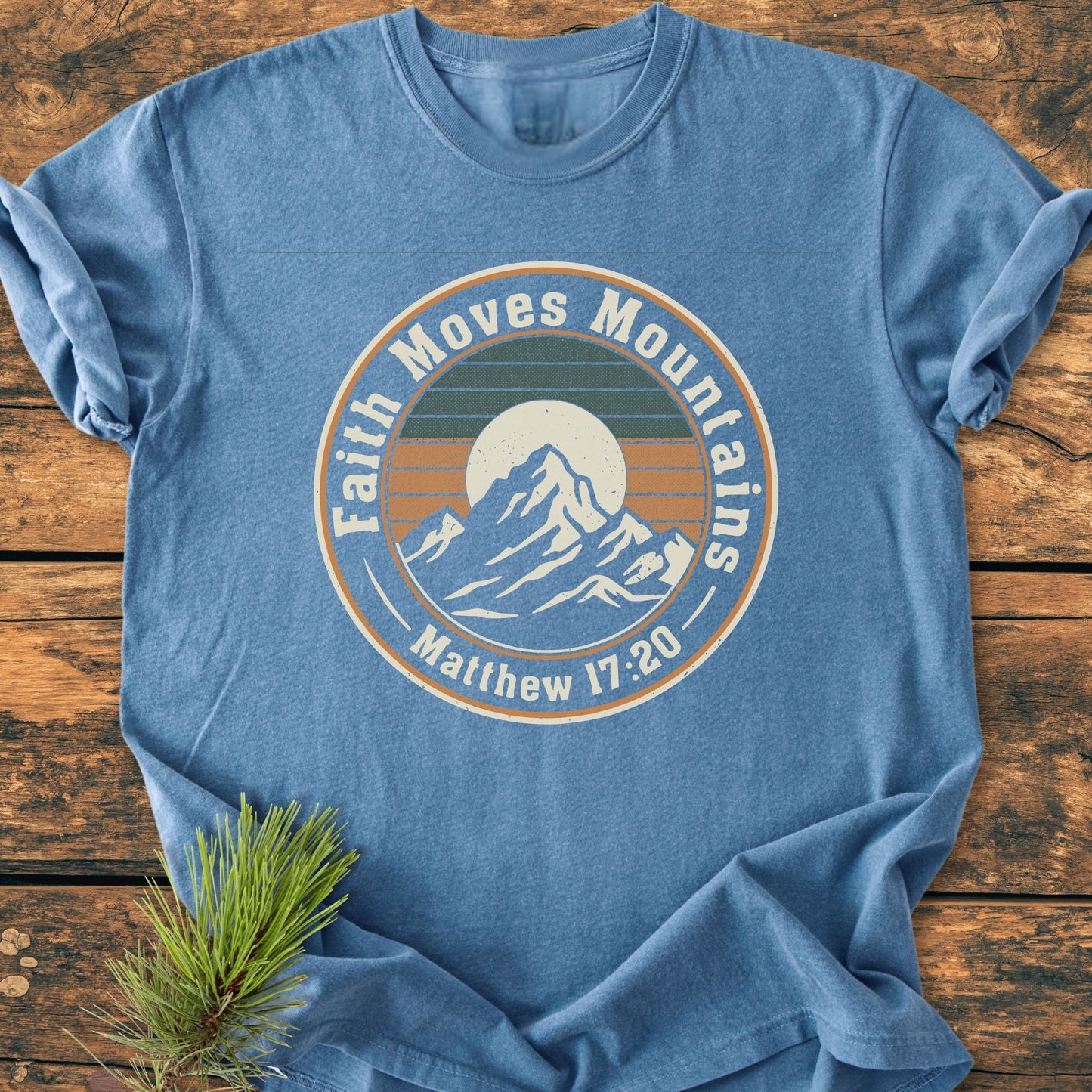 Faith Moves Mountains - Vintage Wash Tee features a round badge graphic with mountains and sun, the phrase Faith Moves Mountains, and Matthew 17:20 on a heather-blue, soft-washed cotton T-shirt.