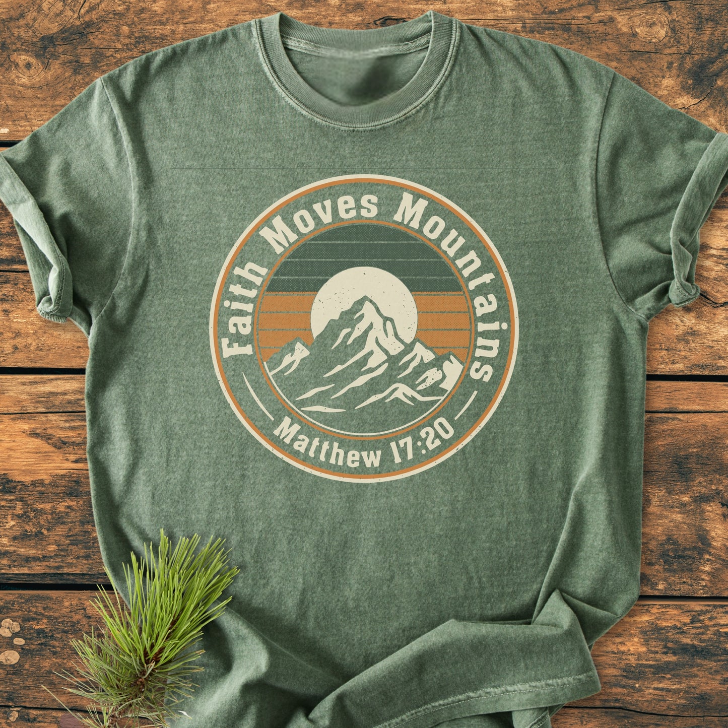 Faith Moves Mountains - Vintage Wash Tee in heather-green, featuring a retro mountain range graphic with a sun and “Matthew 17:20” text, laid flat on a wooden surface with a pine sprig.