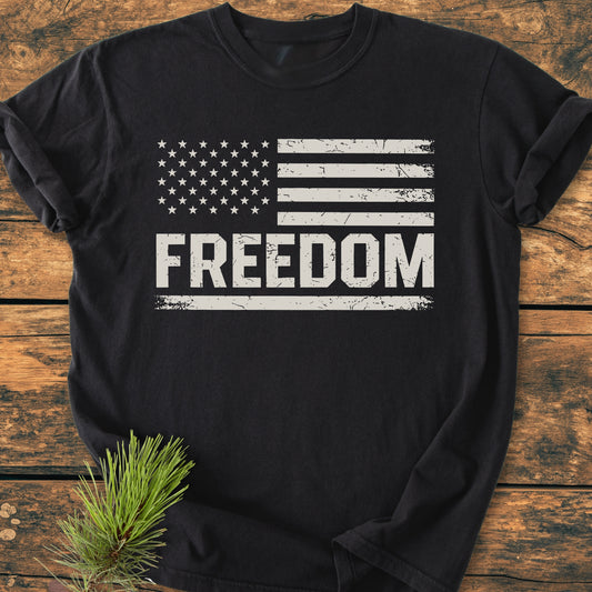 The Freedom Flag - Vintage Wash Tee features a distressed U.S. flag above FREEDOM on a black T-shirt, laid flat on wood with a pine sprig beside it, highlighting its rustic appeal.