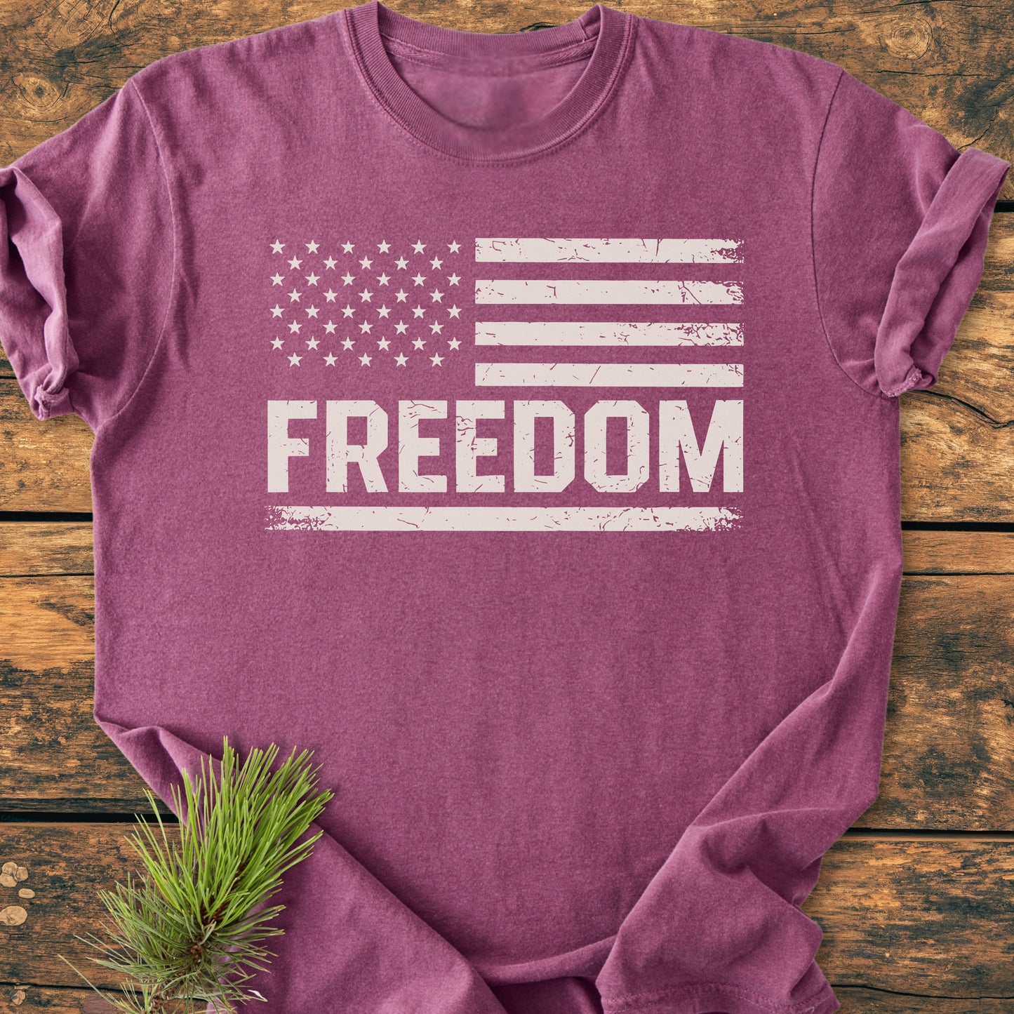 Freedom Flag - Vintage Wash Tee: Heather-purple T-shirt with a distressed white American flag and FREEDOM print, styled with a pine sprig, flat-laid on a wooden surface.